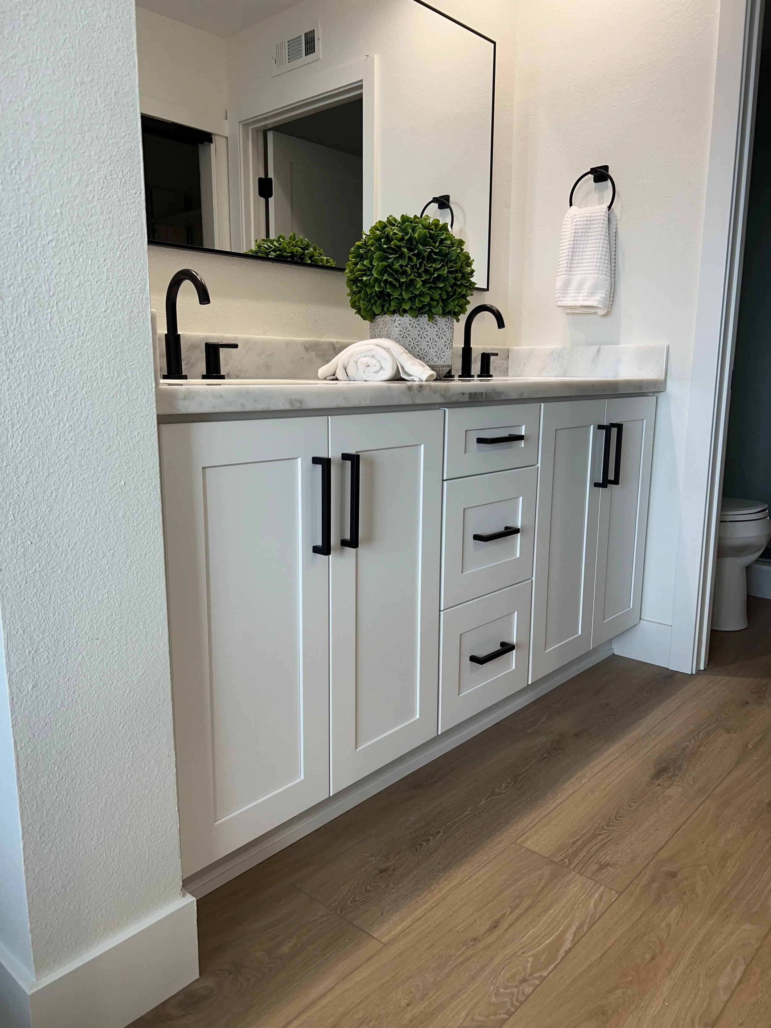 Modern bathroom vanity with white cabinets, black hardware, a marble countertop, a large mirror, a black faucet, a potted green plant, a rolled white towel, and a white hand towel hanging on a black ring.