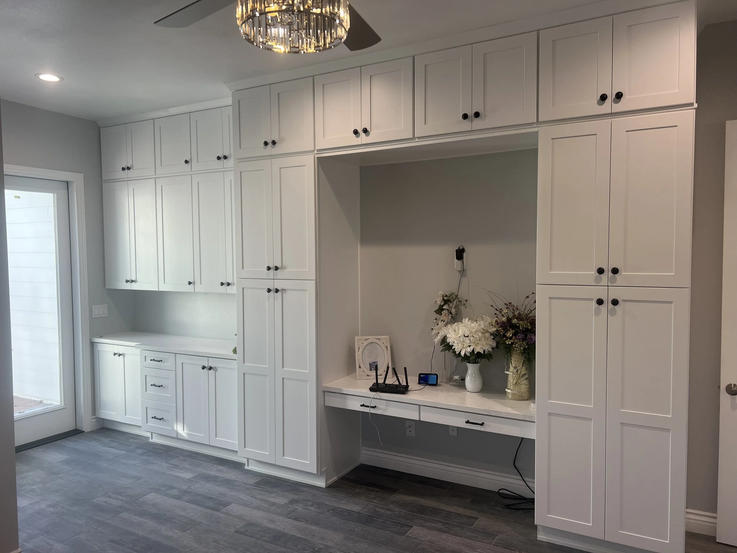 A modern kitchen with tall white cabinets with black knobs, a white countertop, and a decorative vase with flowers. There is a ceiling fan with a light fixture, a window with a view, and a closed door.