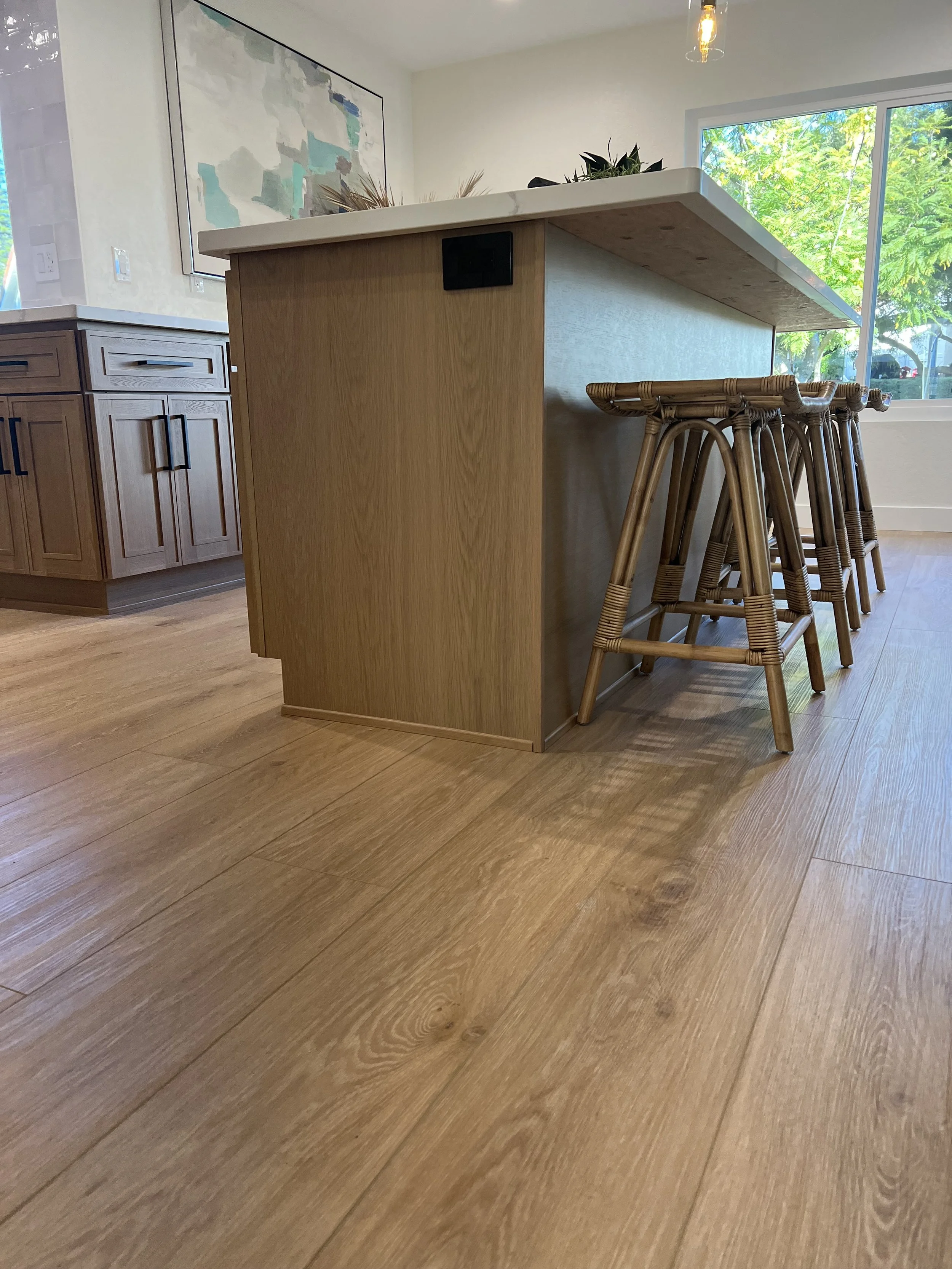 A kitchen island with a marble countertop and wooden base, surrounded by wooden bar stools, with a large window showing green trees outside.
