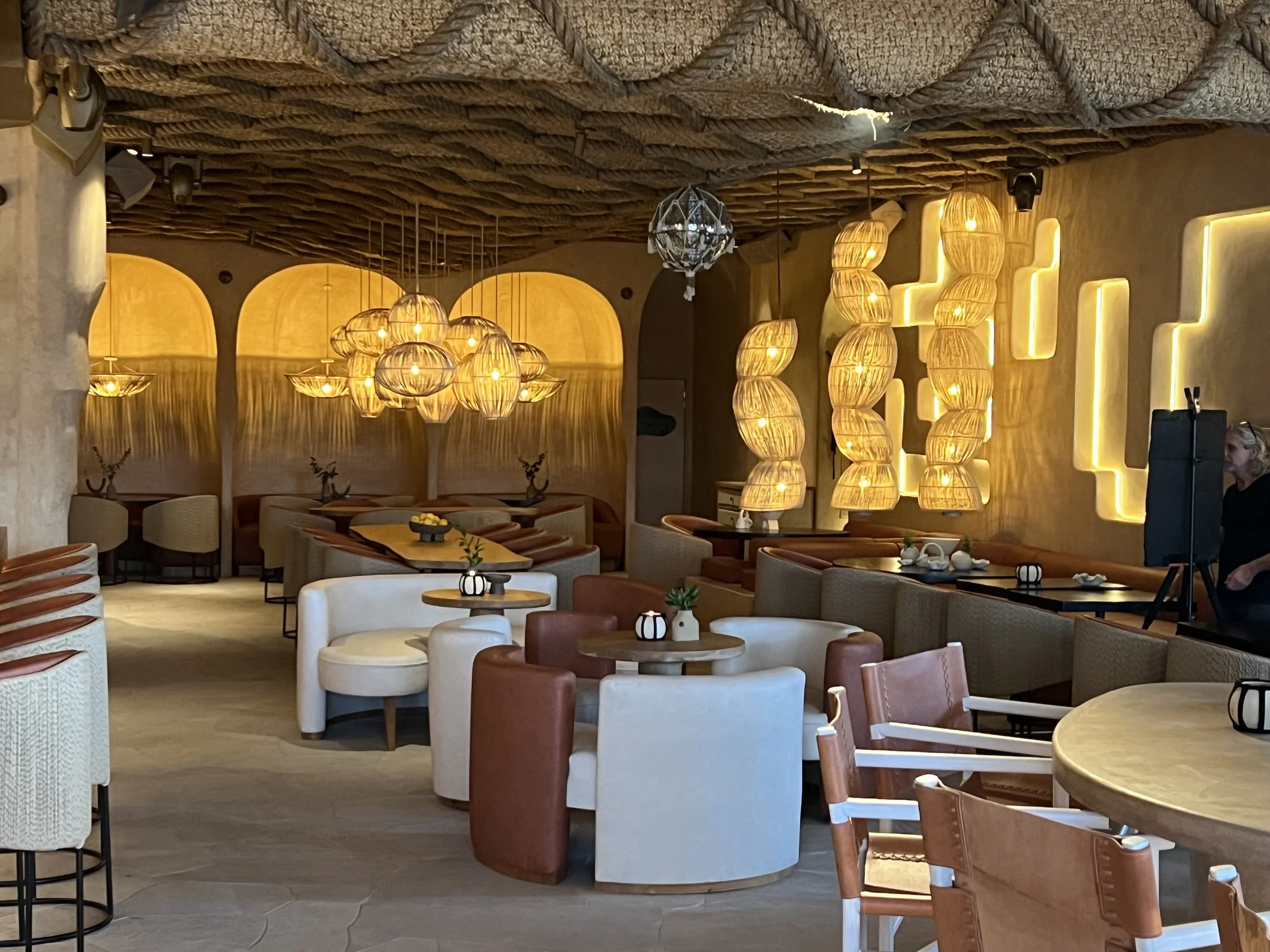 Interior of a restaurant with various chairs and tables, hanging woven light fixtures, a textured ceiling, and warm lighting.