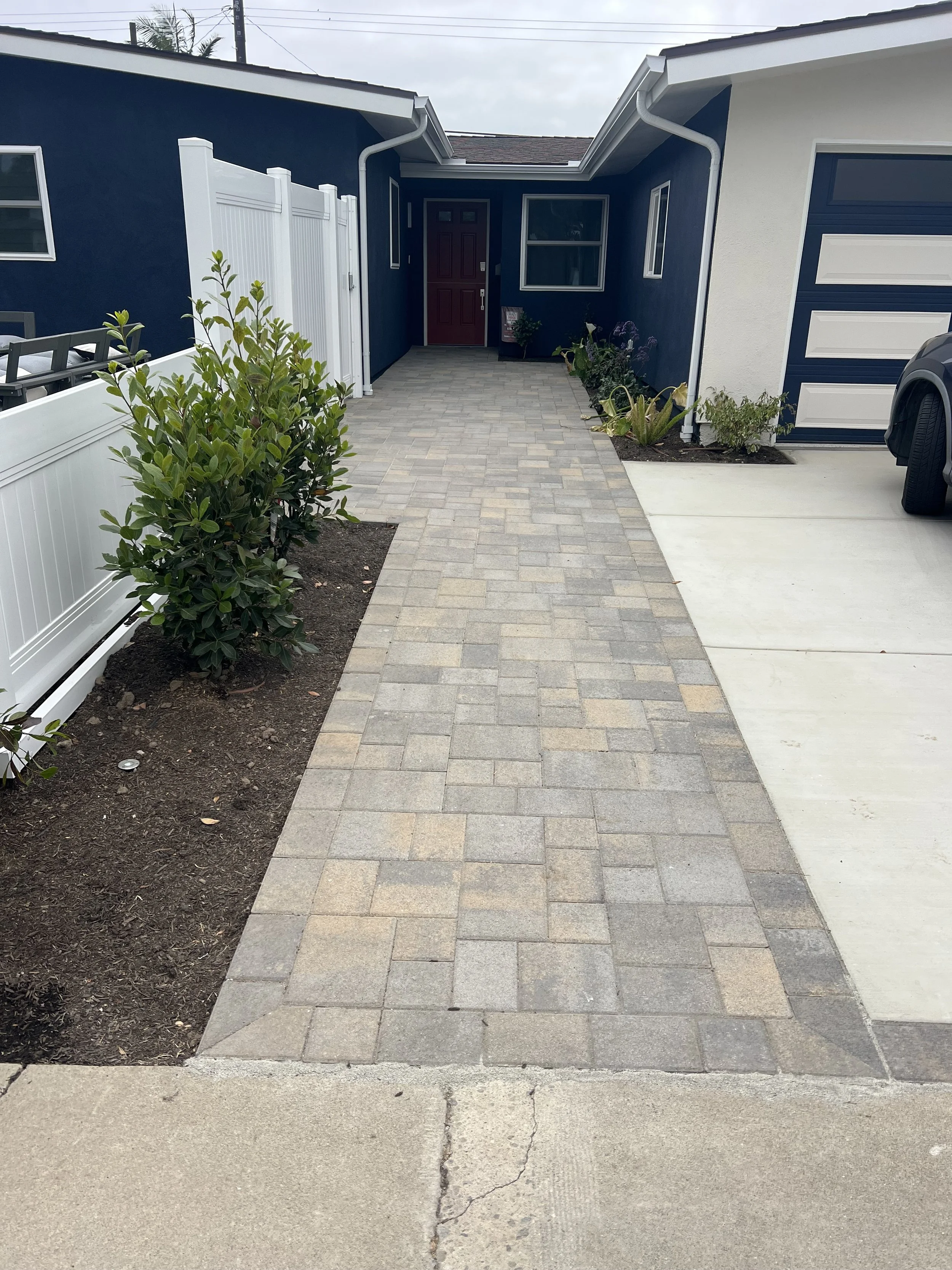 A residential street scene with a driveway, a house with white and blue walls, a garage door, and some plants along the side.