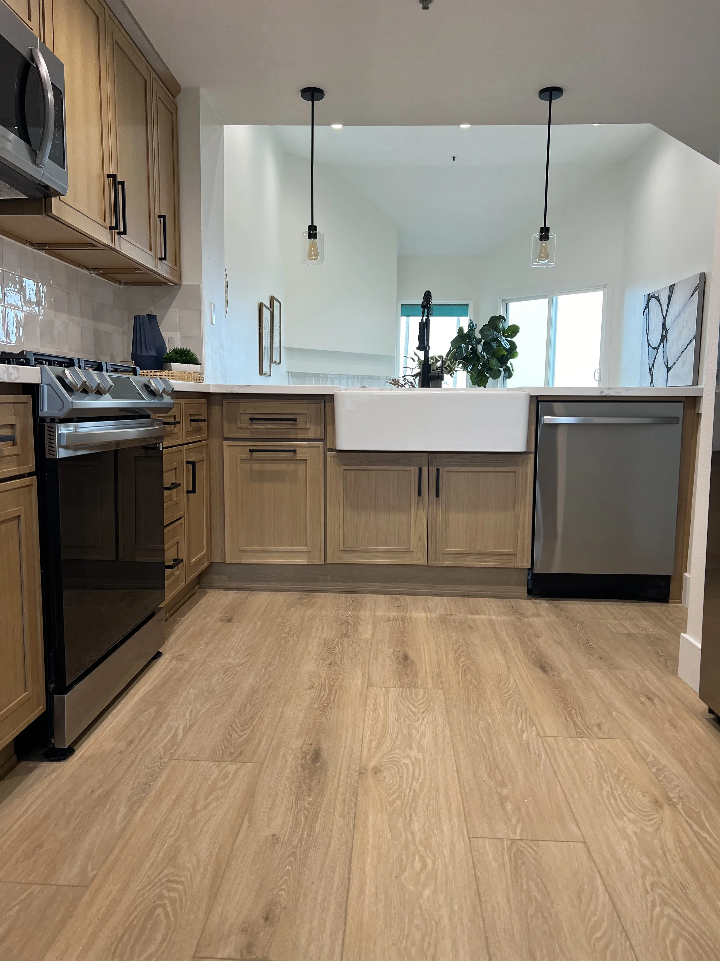 A kitchen with wooden cabinets, a white farmhouse sink, and stainless steel appliances. There are two pendant lights hanging from the ceiling, wooden flooring, and a window in the background with a plant beside it.