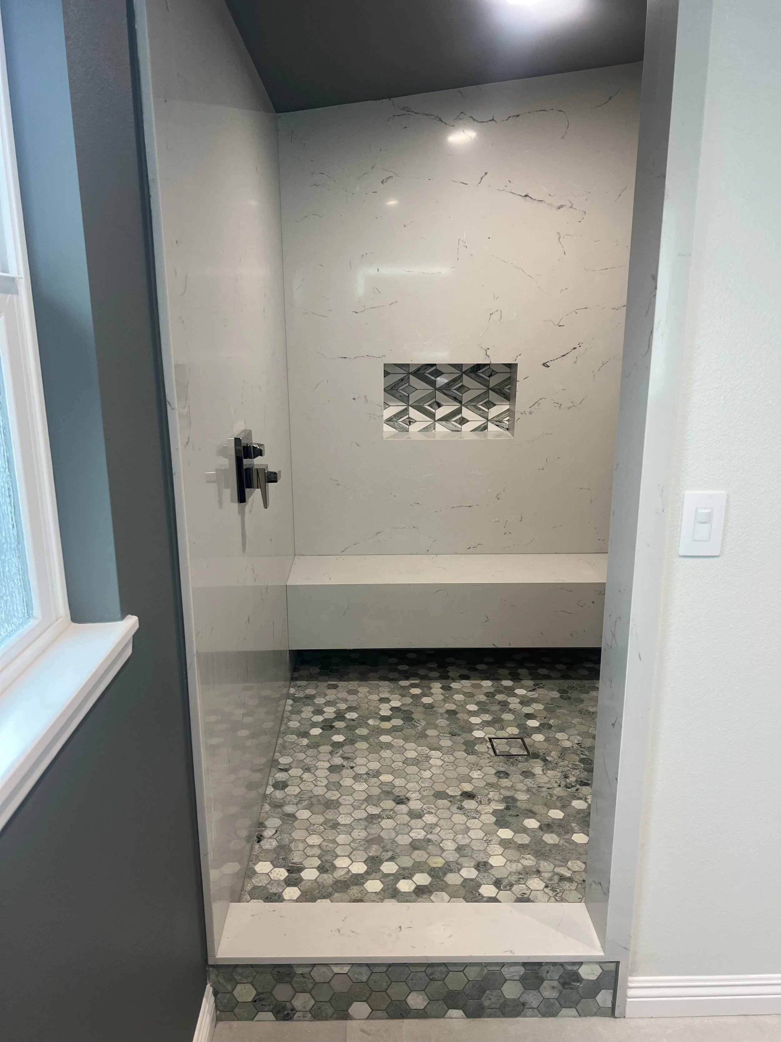 A walk-in shower with a marble wall, a built-in niche with geometric tiles, a matte black shower handle, and a pebble tile floor.