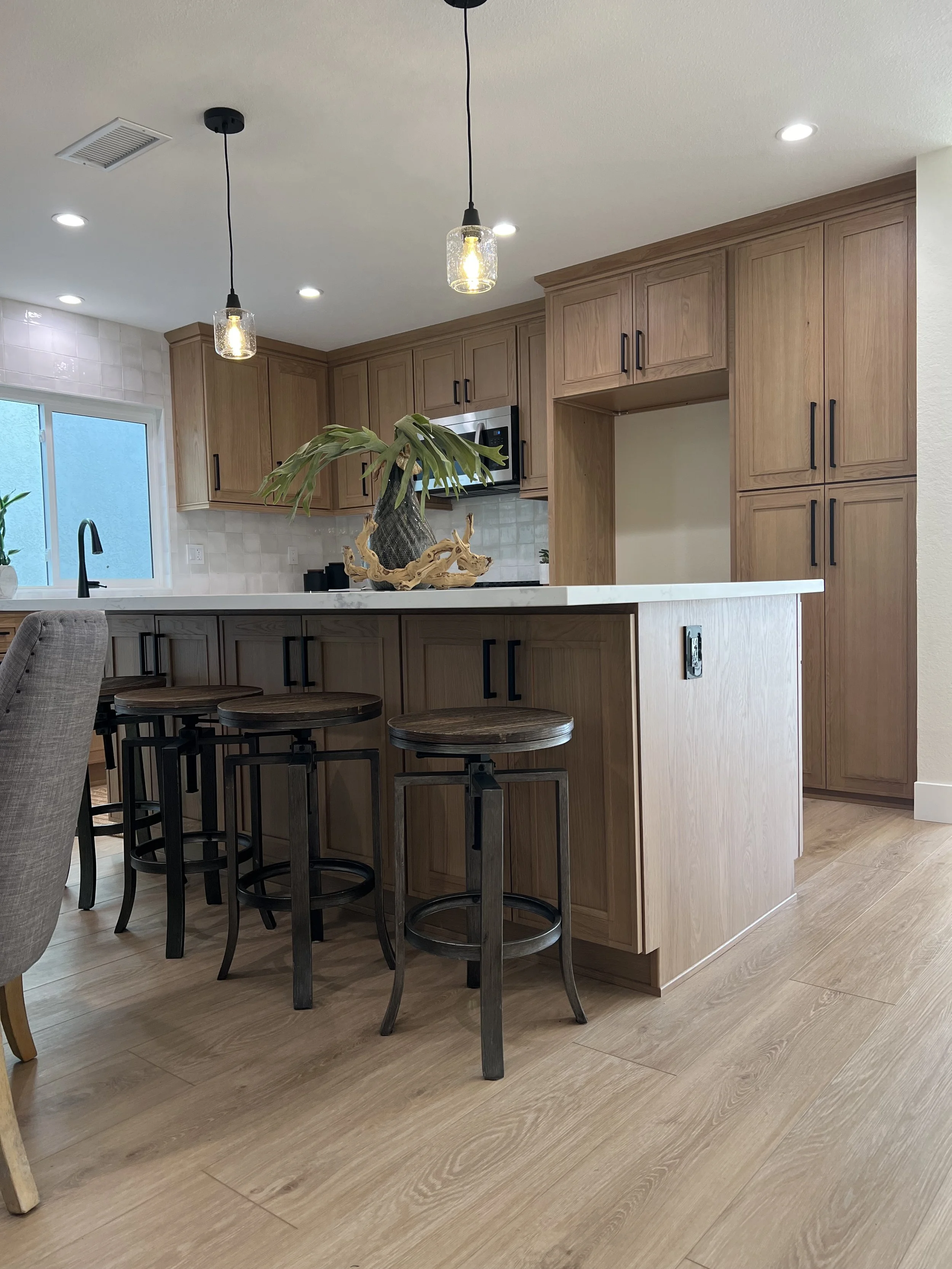 A modern kitchen with wooden cabinets, a white countertop, a black faucet, and a decorative plant on the island. There are black bar stools and a window with frosted glass, with recessed ceiling lights and hanging pendant lights.