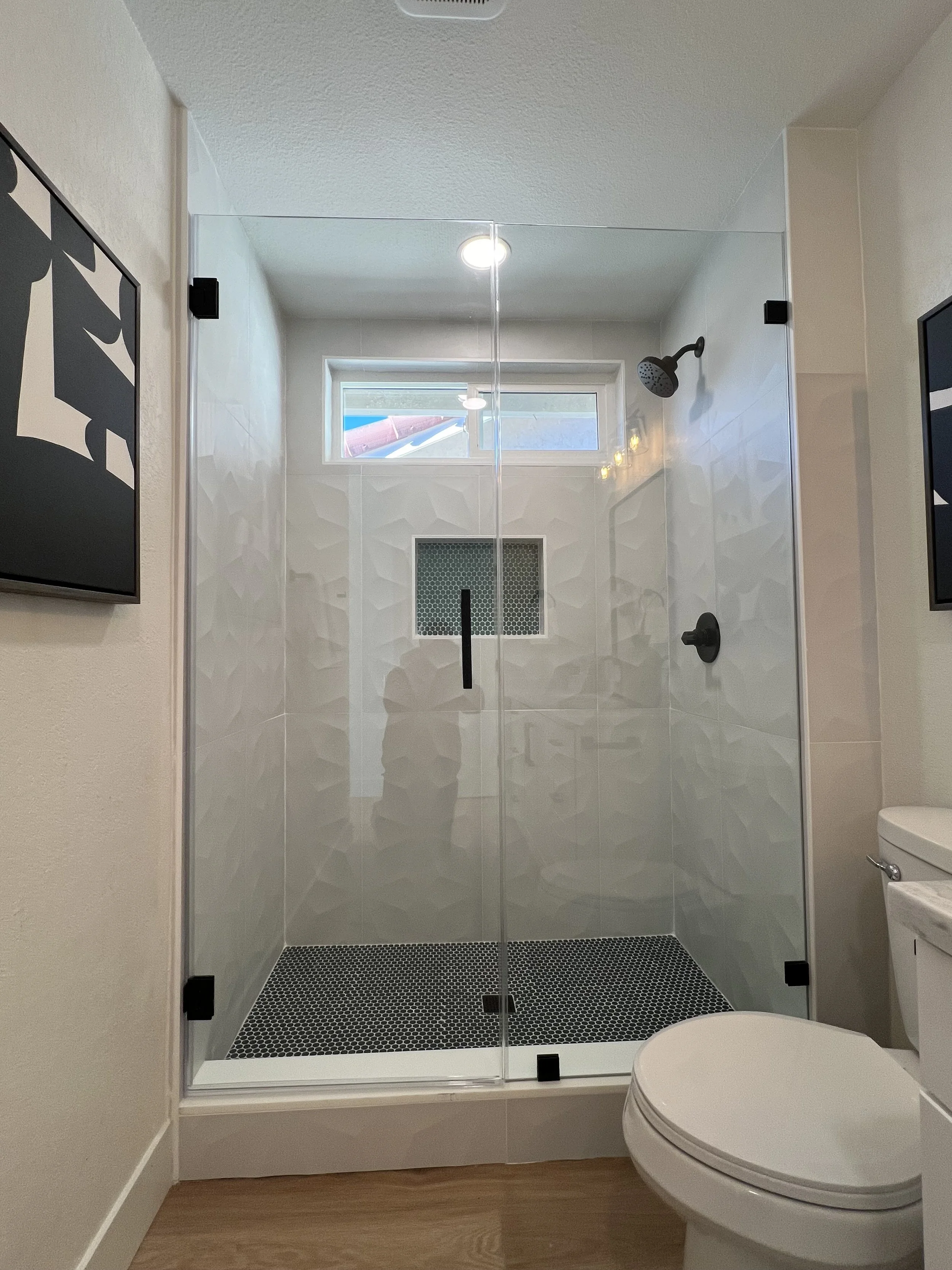 A modern bathroom with a glass-enclosed shower with a black showerhead and handle, a small window, a white toilet to the right, and framed abstract artwork on the left wall.