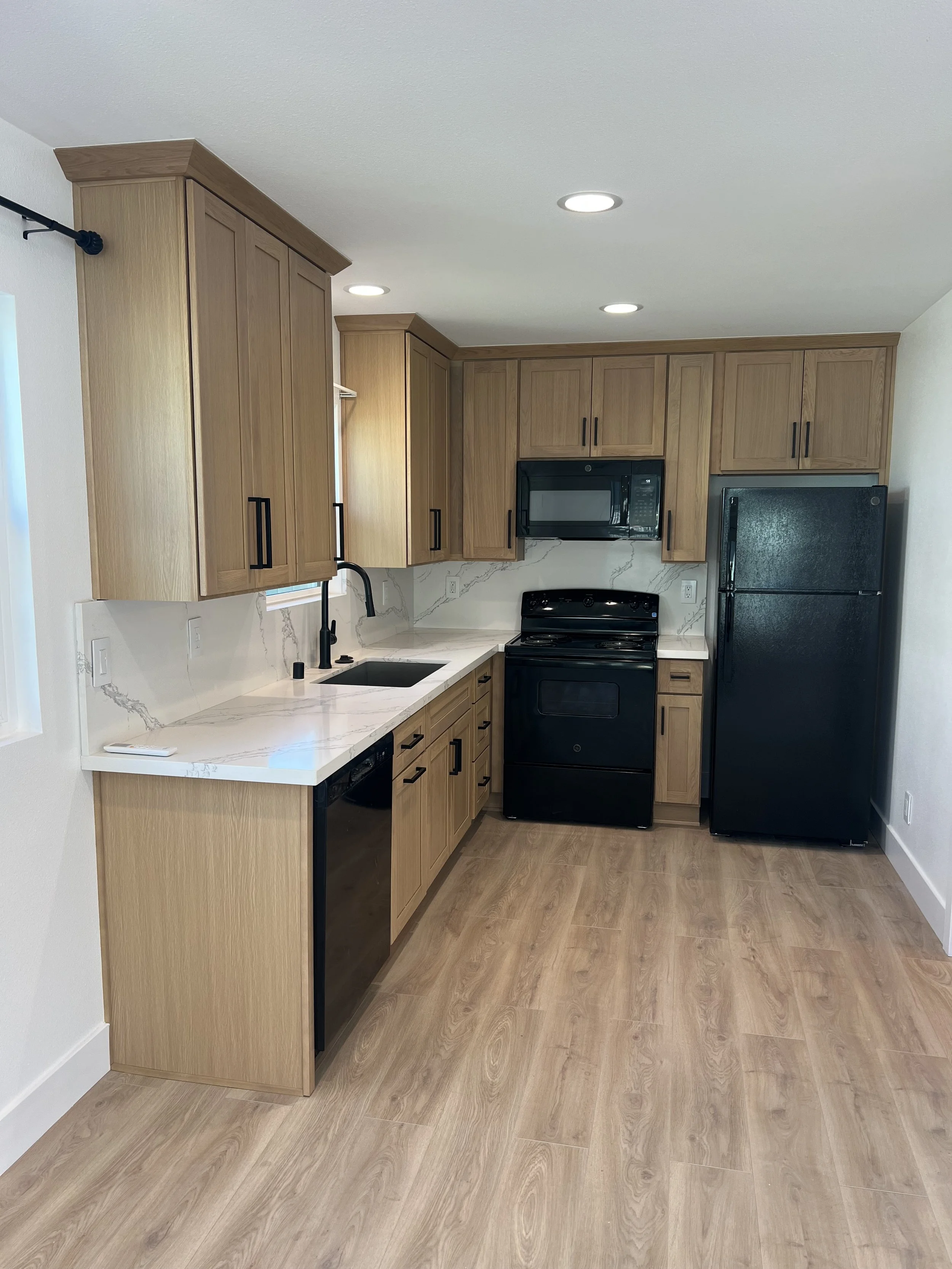 Kitchen with light wood cabinets, white marble countertops, black appliances including a stove, microwave, and refrigerator, and wood flooring.