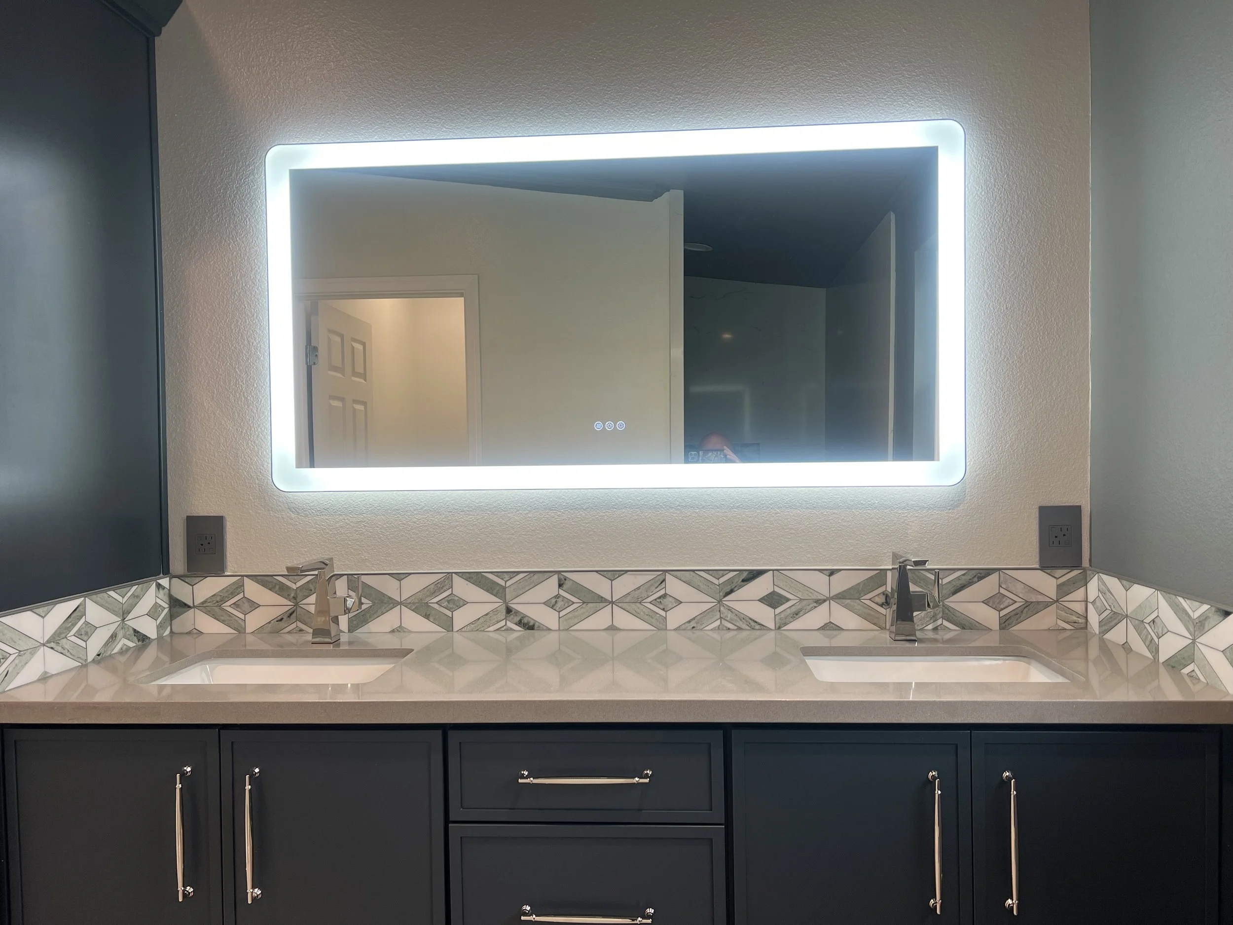 Bathroom vanity with double sinks, dark blue cabinets, silver handles, and a large illuminated mirror with built-in lights, a tiled backsplash with geometric pattern, and part of a countertop visible.