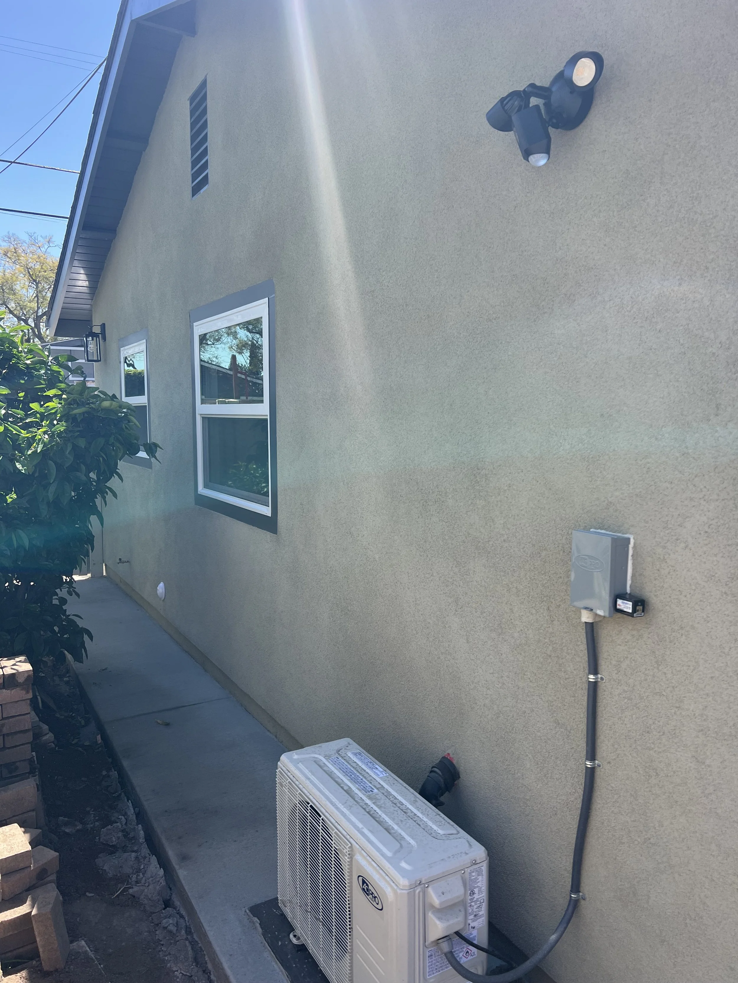Outside wall of a house with two windows, a vent, an outdoor light, an air conditioning unit, and electrical connections.