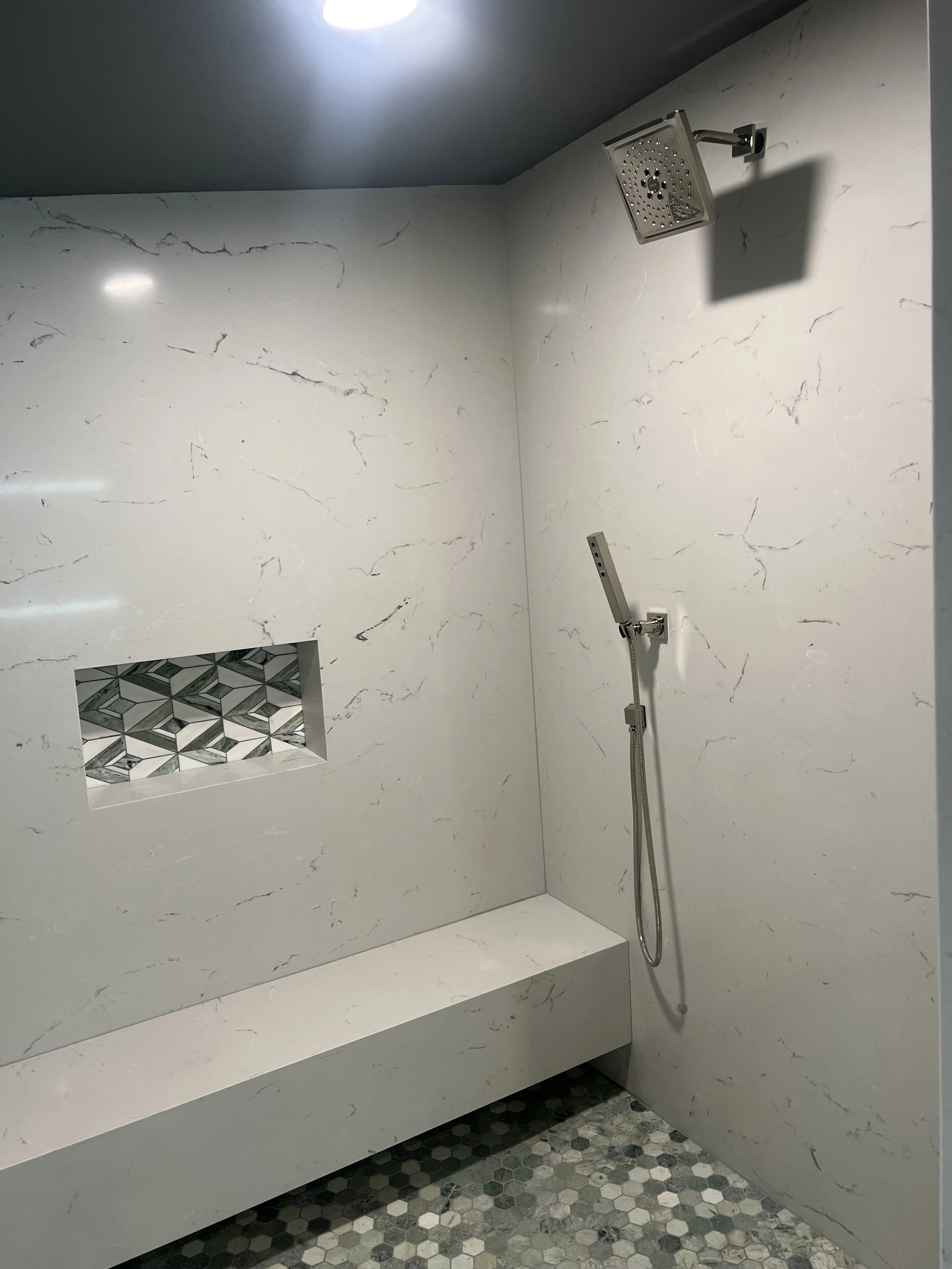 A modern shower with a marble wall, a square rain showerhead, a handheld shower, and a patterned hexagonal tile floor.