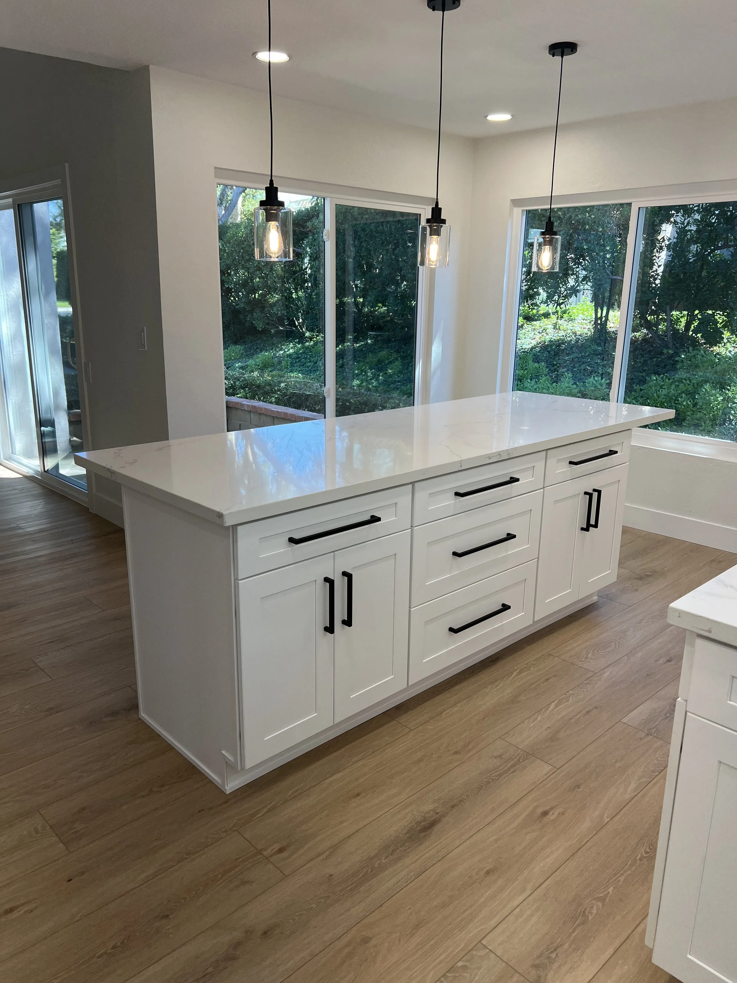Bright kitchen with white island, three pendant lights overhead, large windows with greenery outside, and hardwood floors.