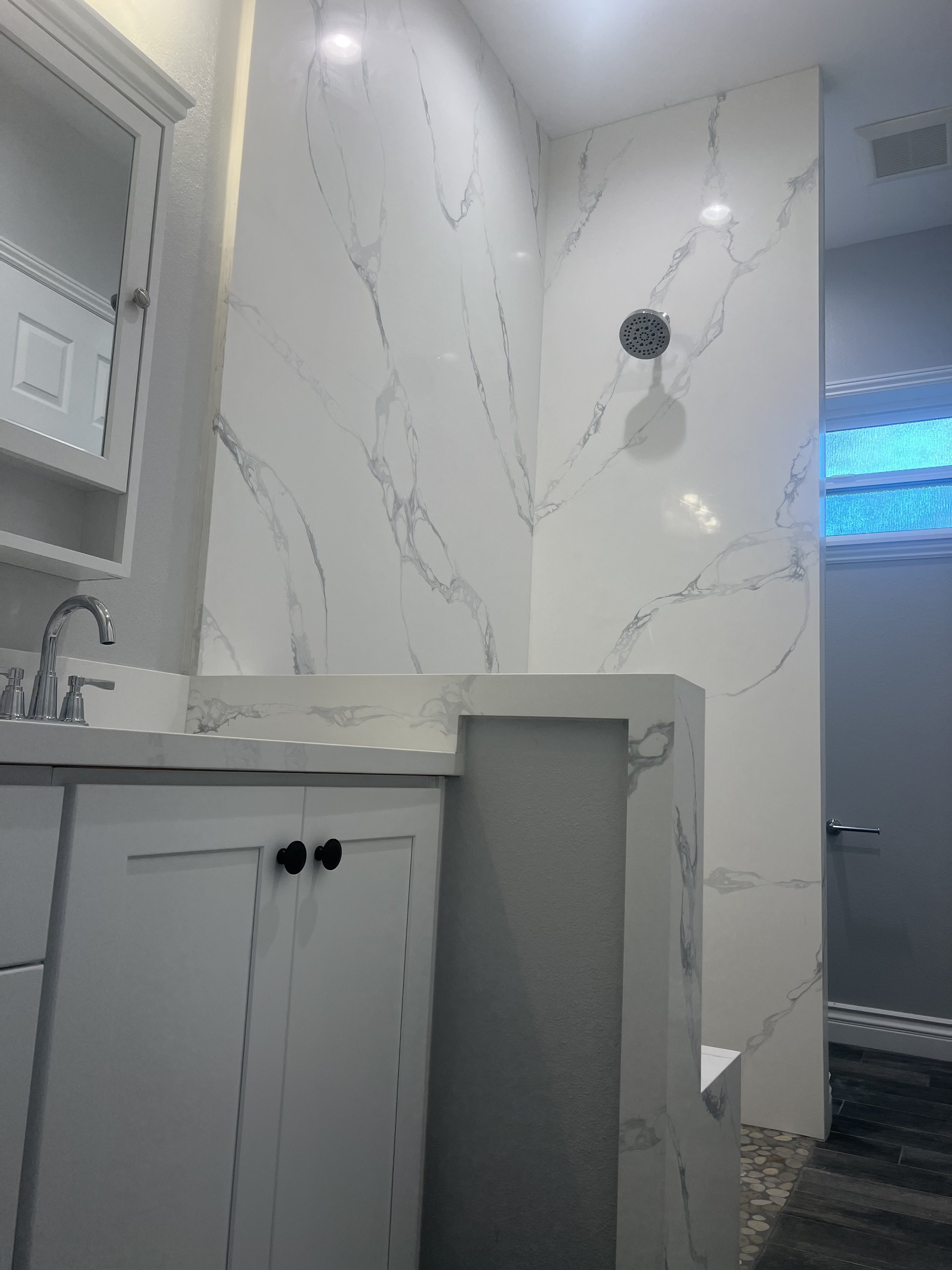 View of a bathroom with white marble walls with gray veining, a white vanity with black knobs, a mirror cabinet, and a drain. The floor has a pebble mosaic section near the shower, which features a rain showerhead and a glass door with a blue tint.