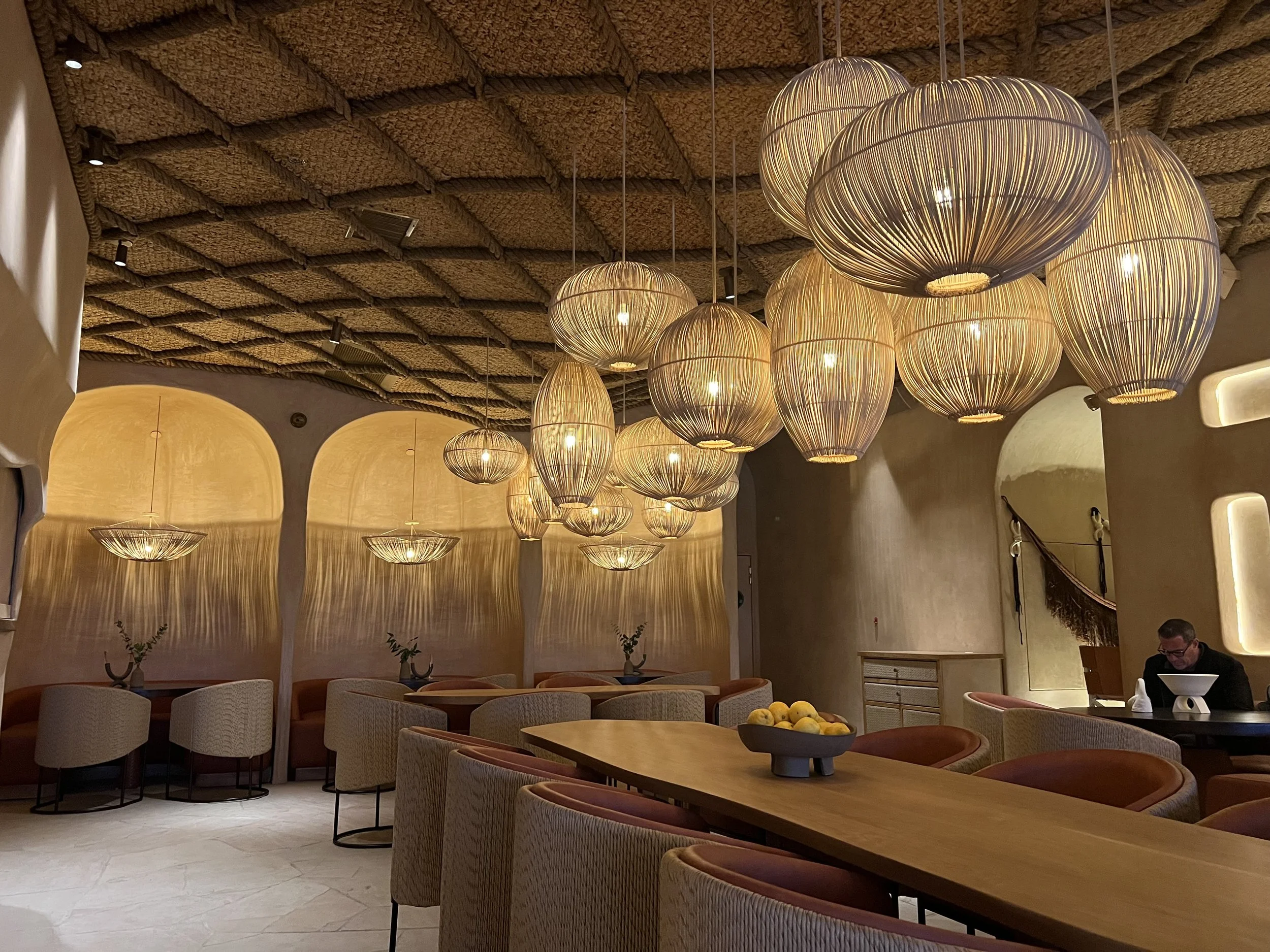 Interior of a restaurant with warm lighting, wooden tables and chairs, modern pendant lights hanging from a textured ceiling, and a person sitting at a table in the corner.