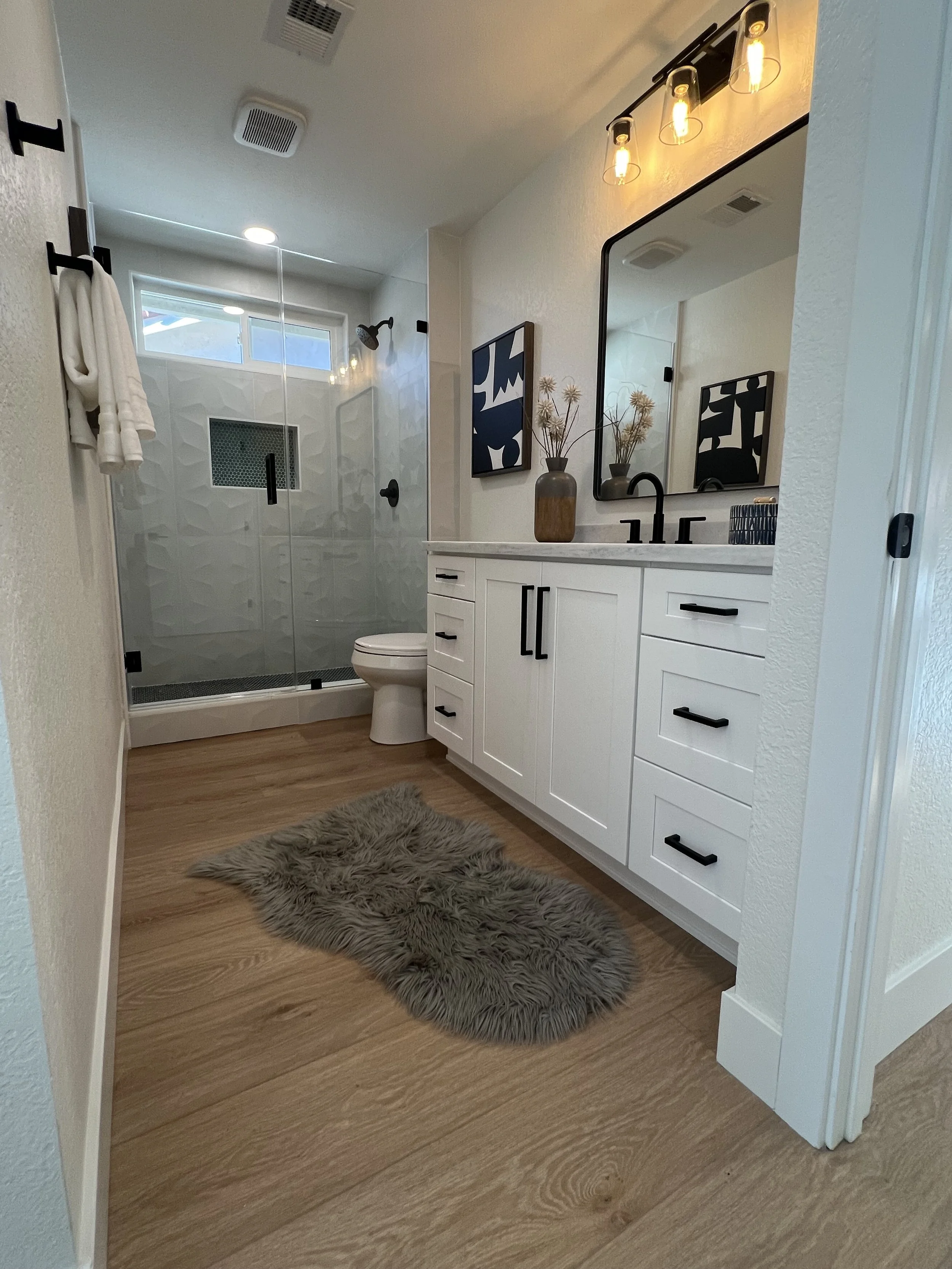 Modern bathroom featuring a glass-enclosed walk-in shower with black fixtures and a window, a white vanity with black handles, a large mirror, framed artwork, a vase with dried flowers, a black soap dispenser, a fluffy gray rug on a wood floor, and o