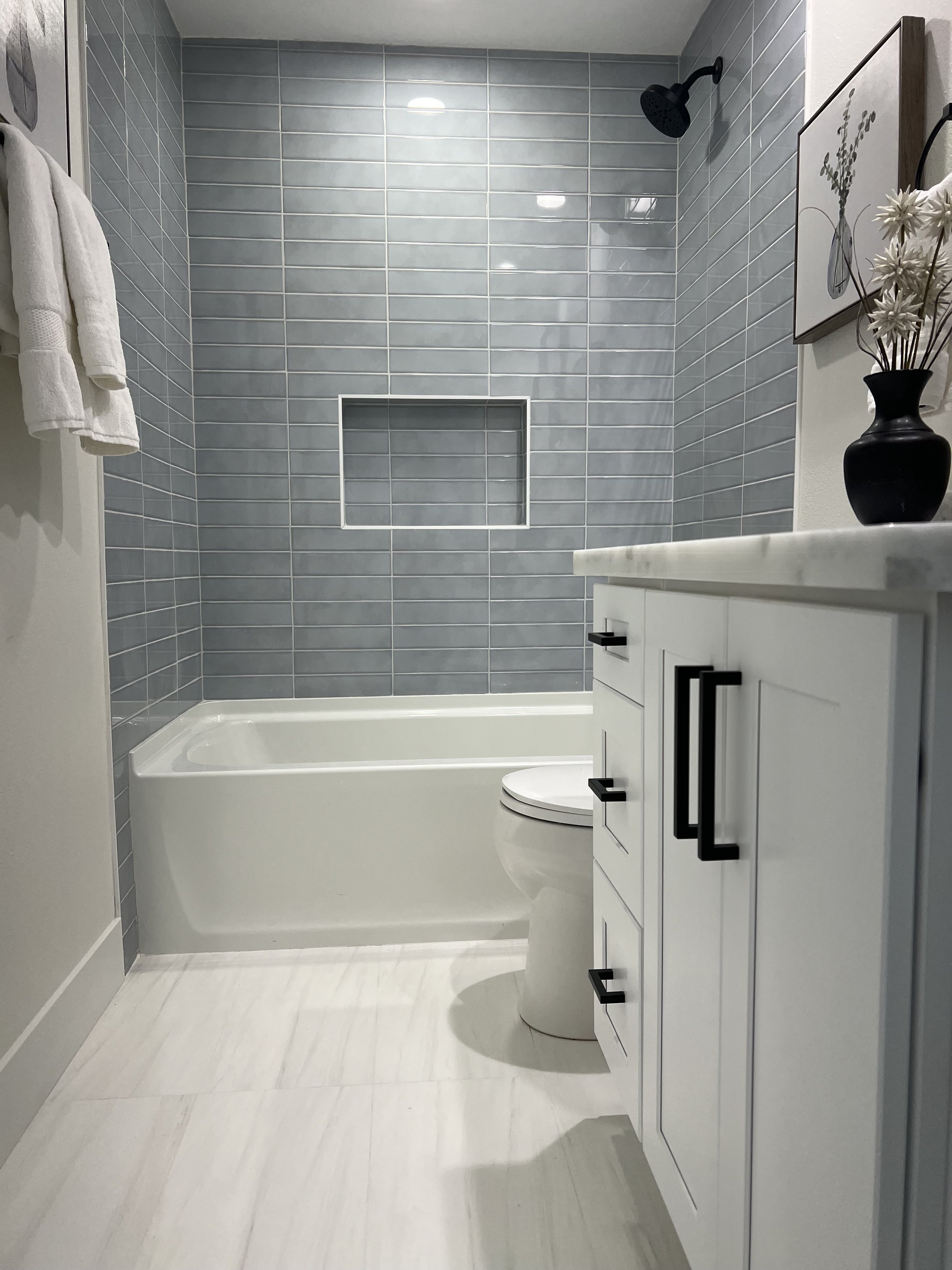 Modern bathroom with blue tiled shower area, white vanity cabinet, black fixtures, and a black vase with white flowers on the counter.