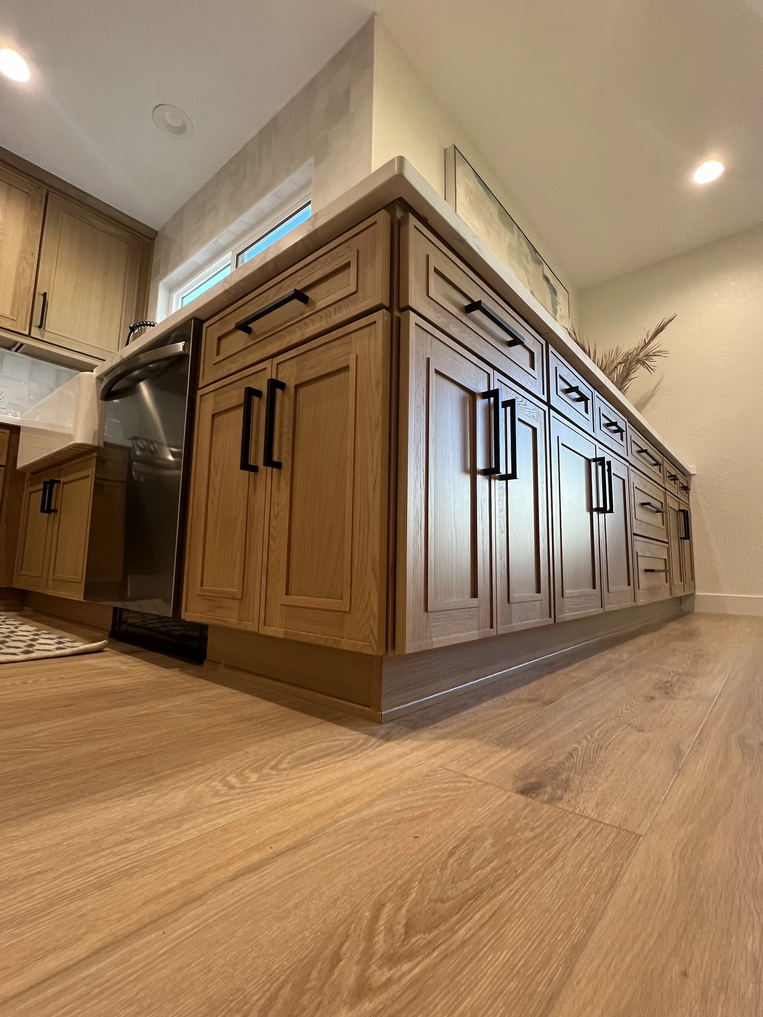 Wooden kitchen cabinets with black handles, a countertop with a plant, hardwood flooring, and a small window behind the counter, with ceiling lights illuminating the space.