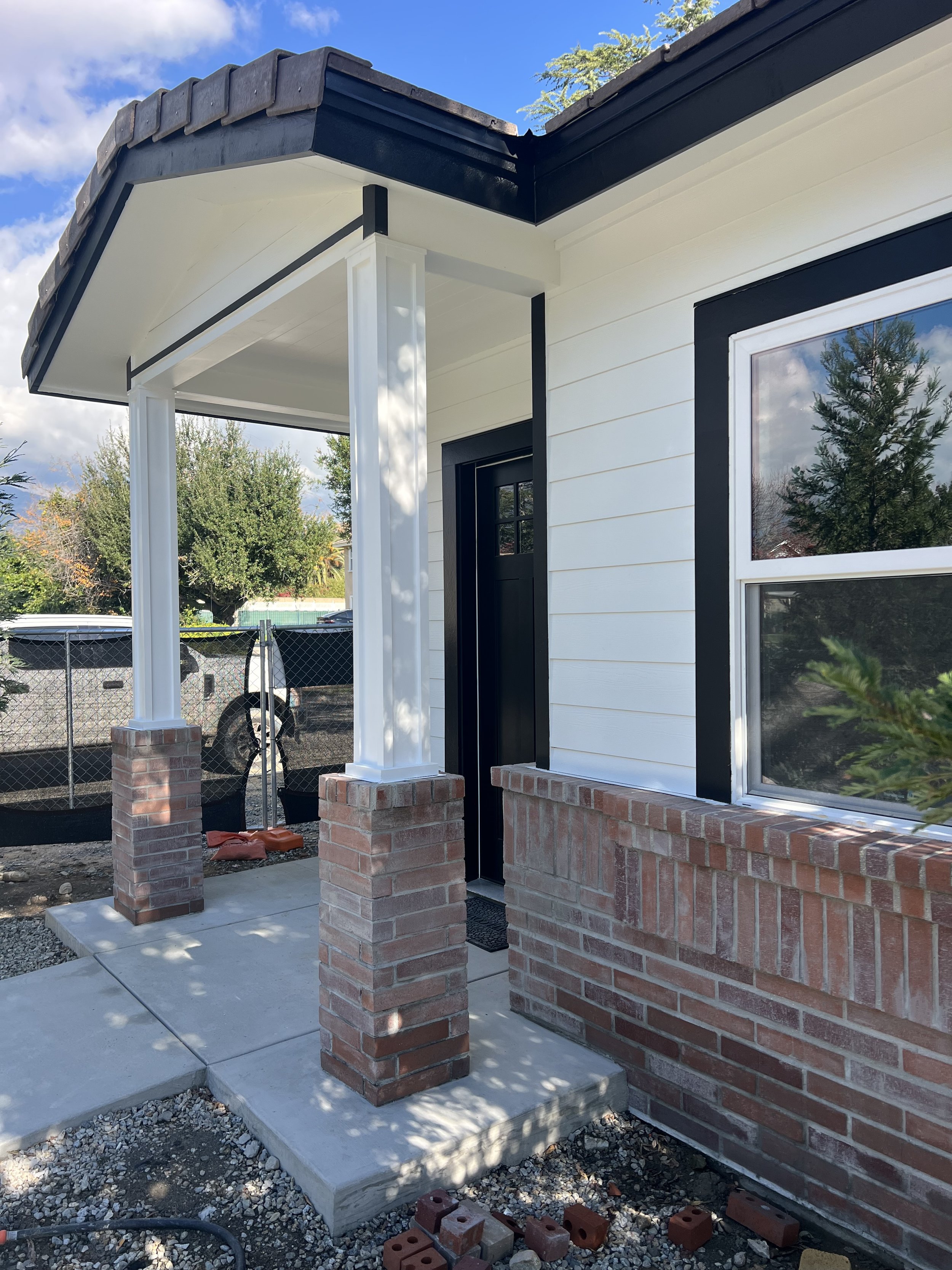 Front porch of a house under construction with brick columns, concrete slab, and a black door, surrounded by gravel and construction materials.