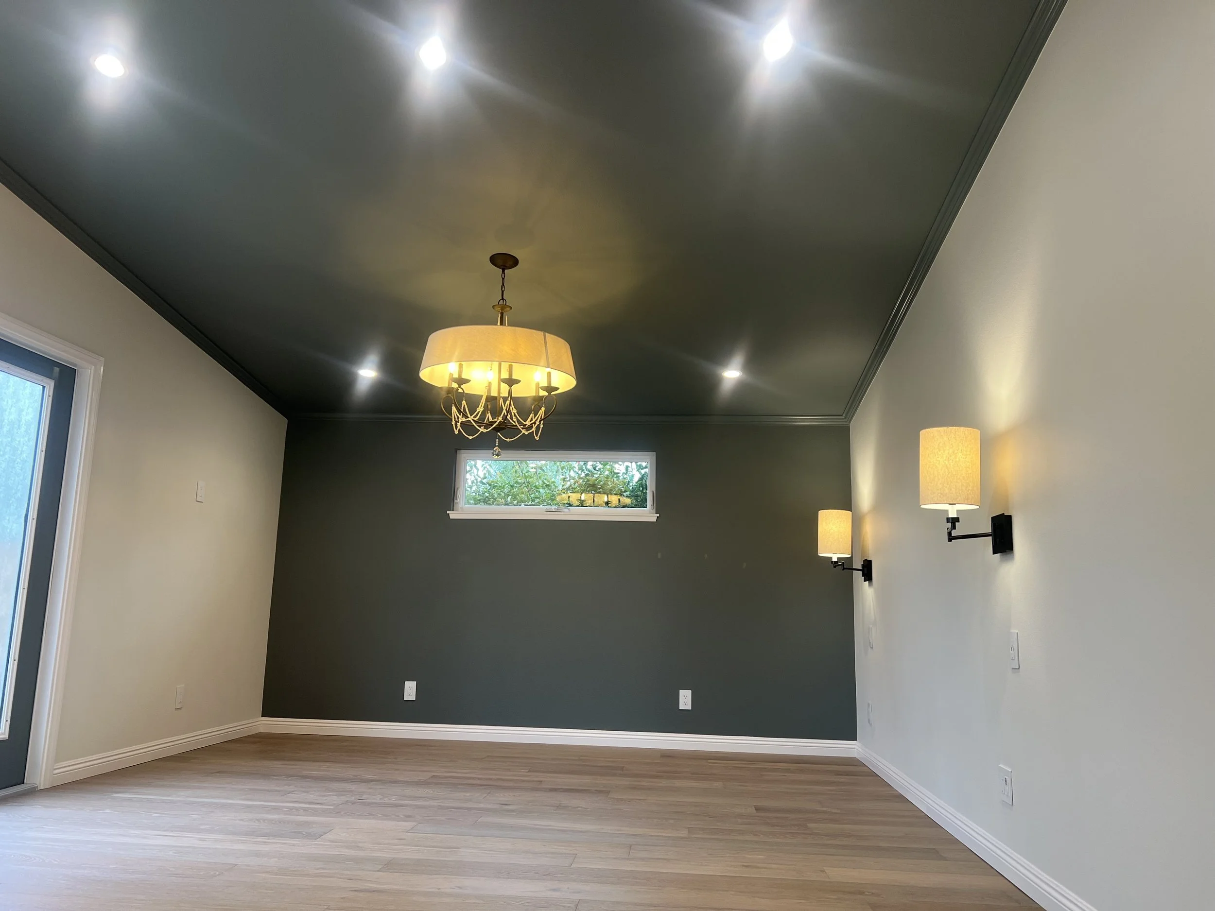 Empty living room with light-colored walls, a dark accent wall, a chandelier, wall-mounted lamps, a window showing greenery outside, and a hardwood floor.