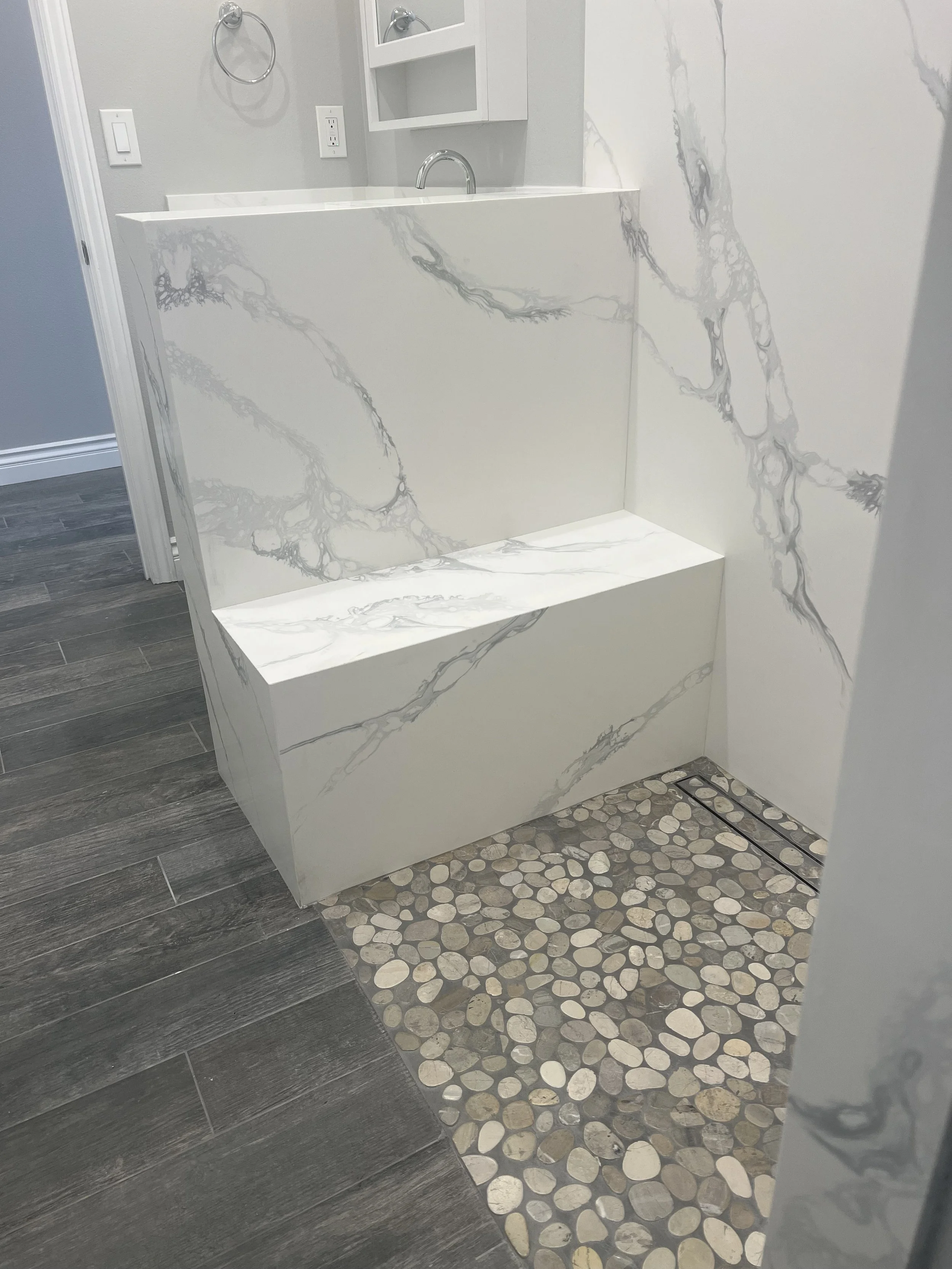 Close-up photo of a bathroom with a marble vanity and a pebble stone shower floor, featuring a white marble wall and dark wood-like floor tiles.