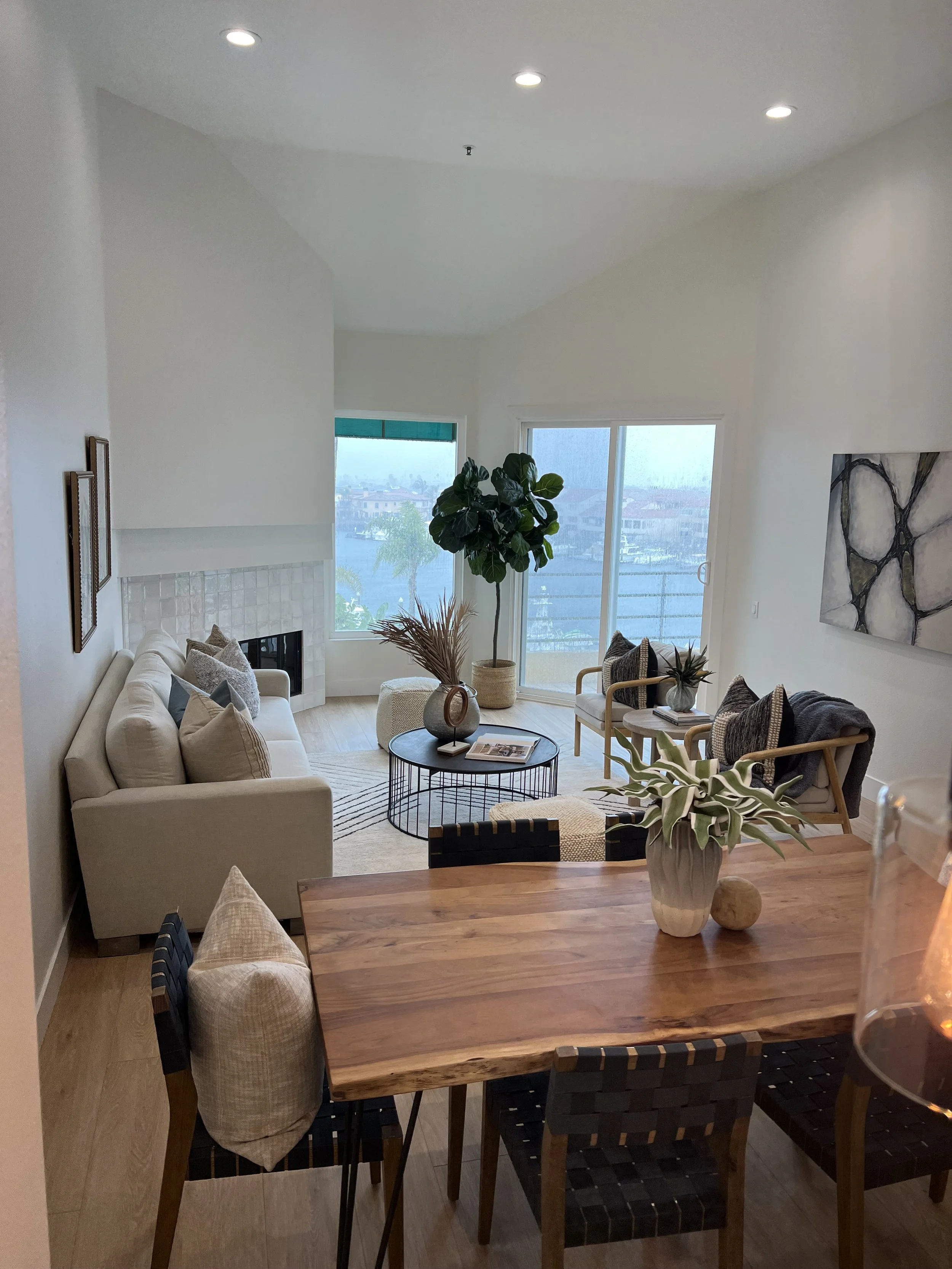 Living room with a beige sofa, wooden coffee table, large potted plant, and framed artwork, with windows showing a view of a waterfront and buildings outside.