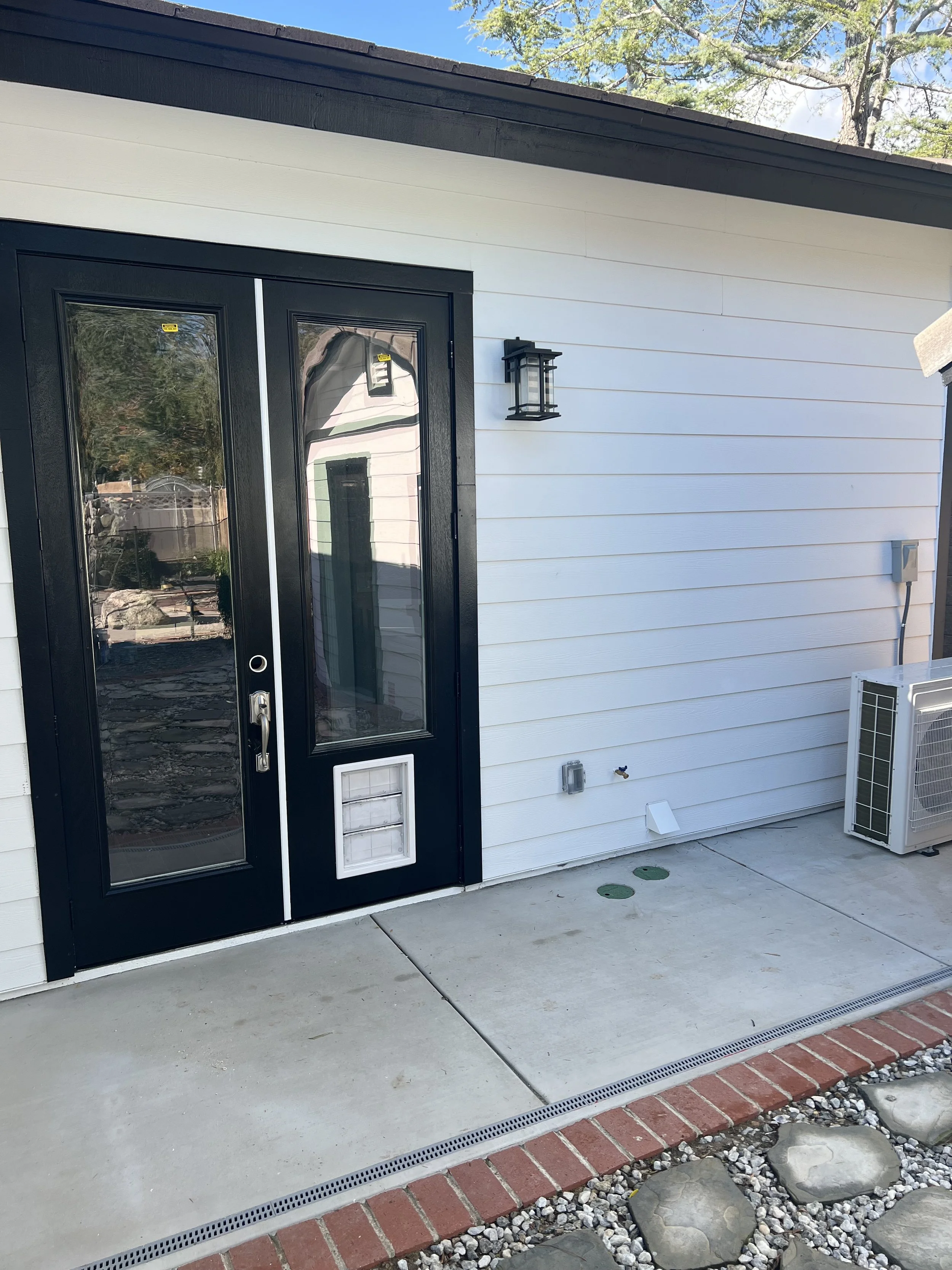 The exterior of a house showing a black front door with glass panels, a wall-mounted outdoor light fixture, a small ventilation opening, and an air conditioning unit nearby. The house has white horizontal siding, and the porch area has a concrete flo