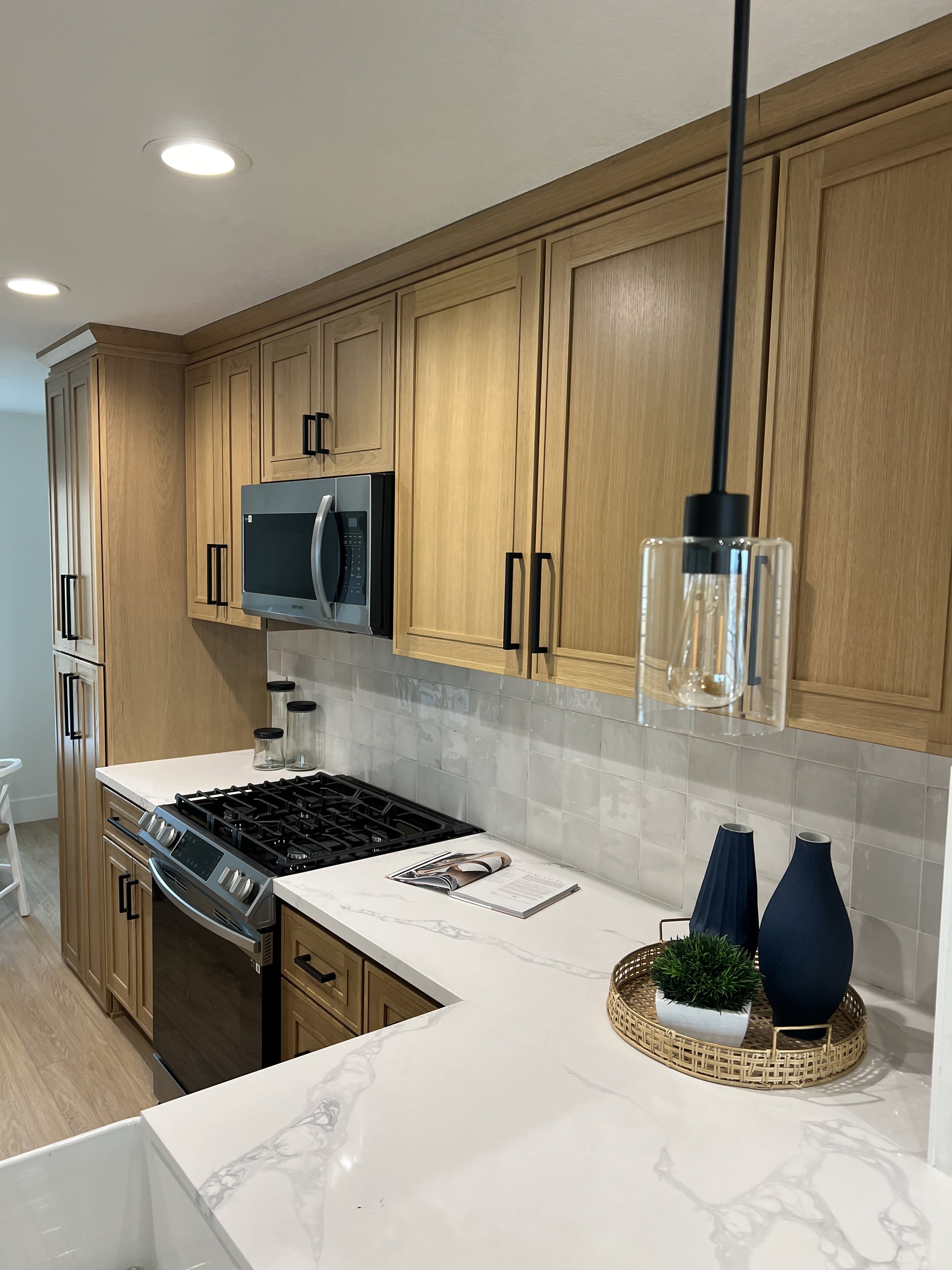 Kitchen with natural wood cabinets, stainless steel microwave and oven, white countertop, decorative vases, and an open cookbook on the counter.