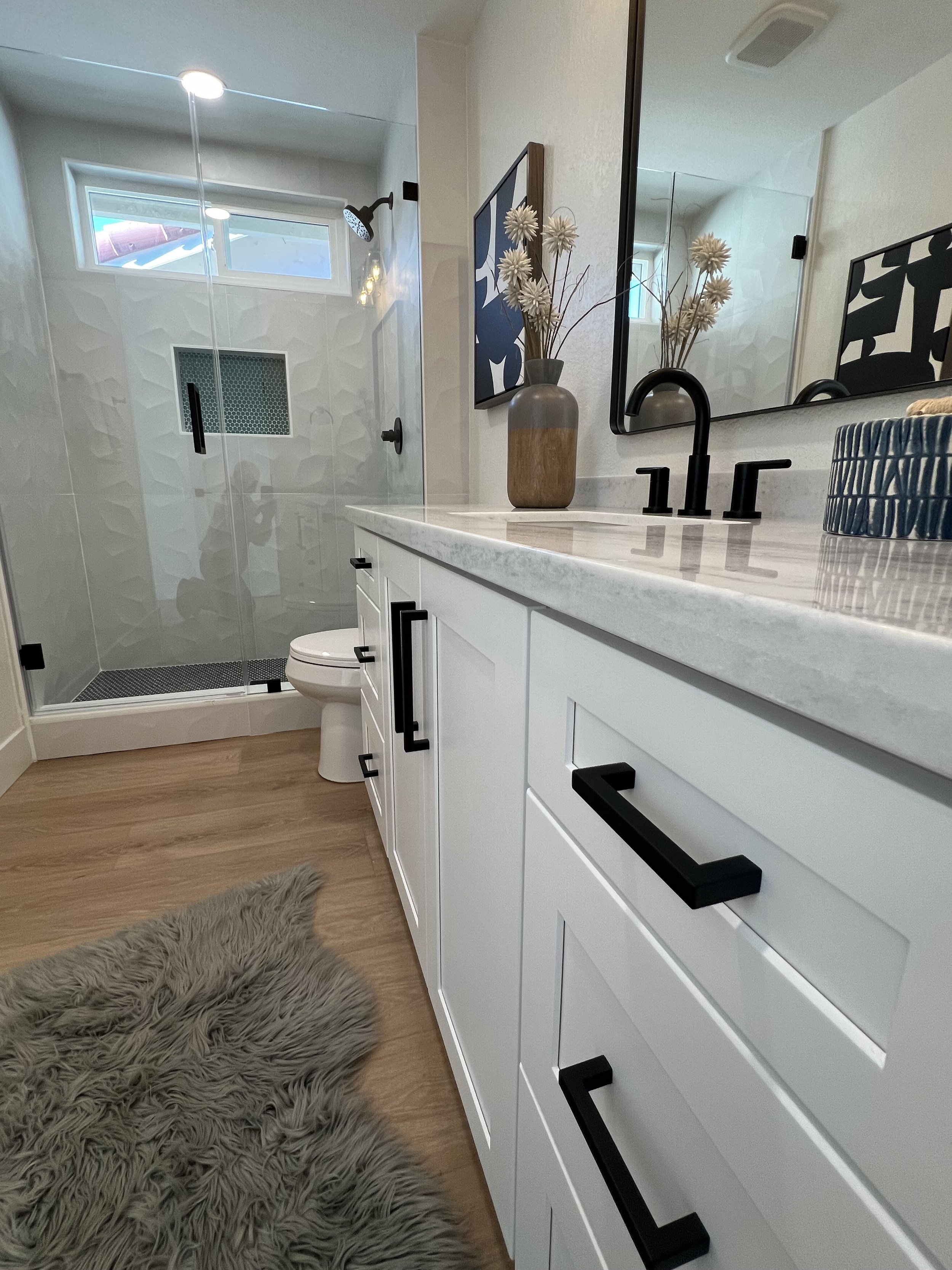 Modern bathroom with walk-in glass shower, white vanity with black handles, black fixtures, decorative vase with flowers, large mirror, wooden flooring, and a fluffy gray rug.