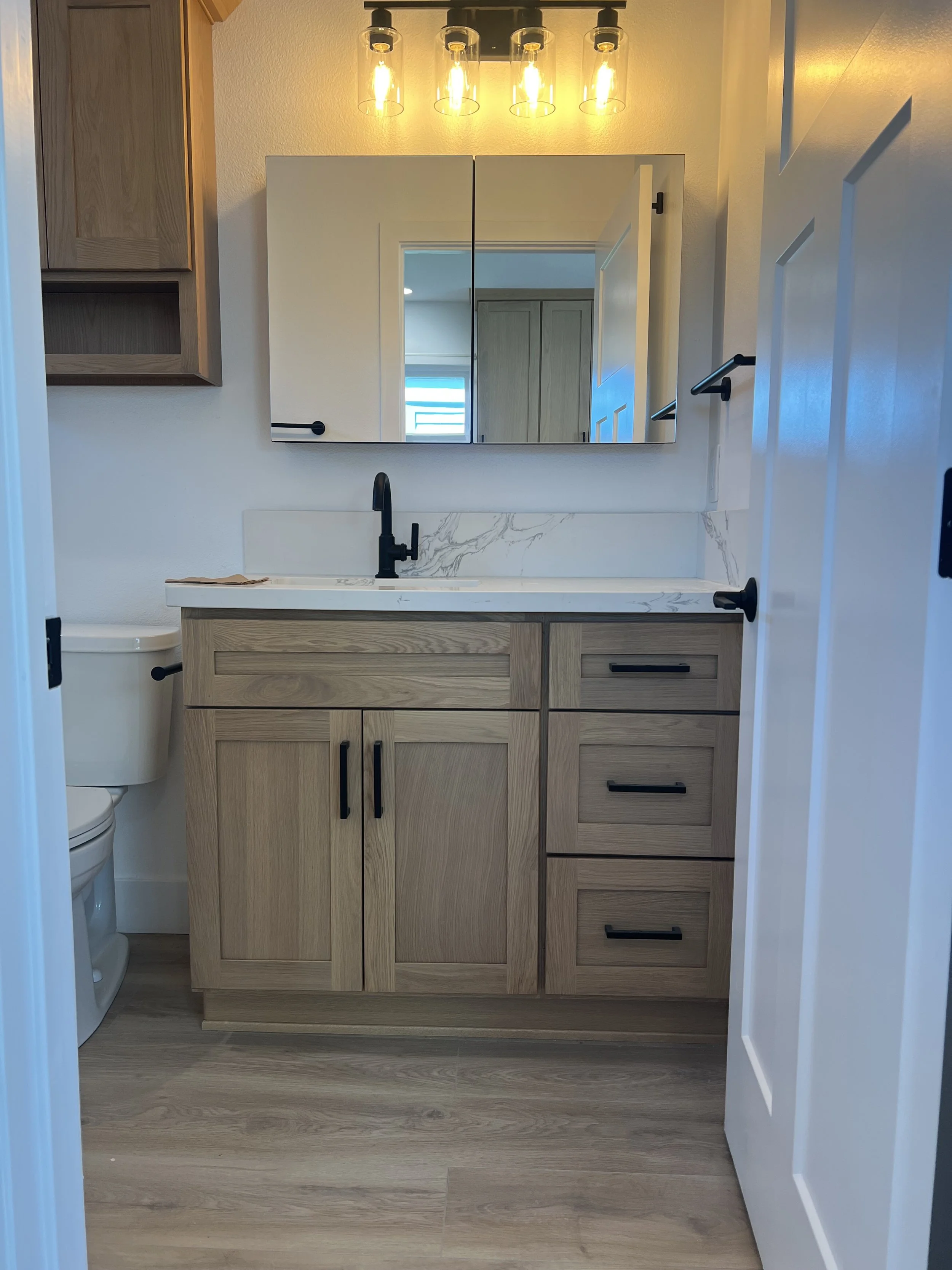 A modern bathroom vanity with a light wooden cabinet, black hardware, and a white marble countertop with gray veining. A black sink faucet is installed on the countertop. A mirror is mounted above the vanity, reflecting a nearby room. To the left, pa