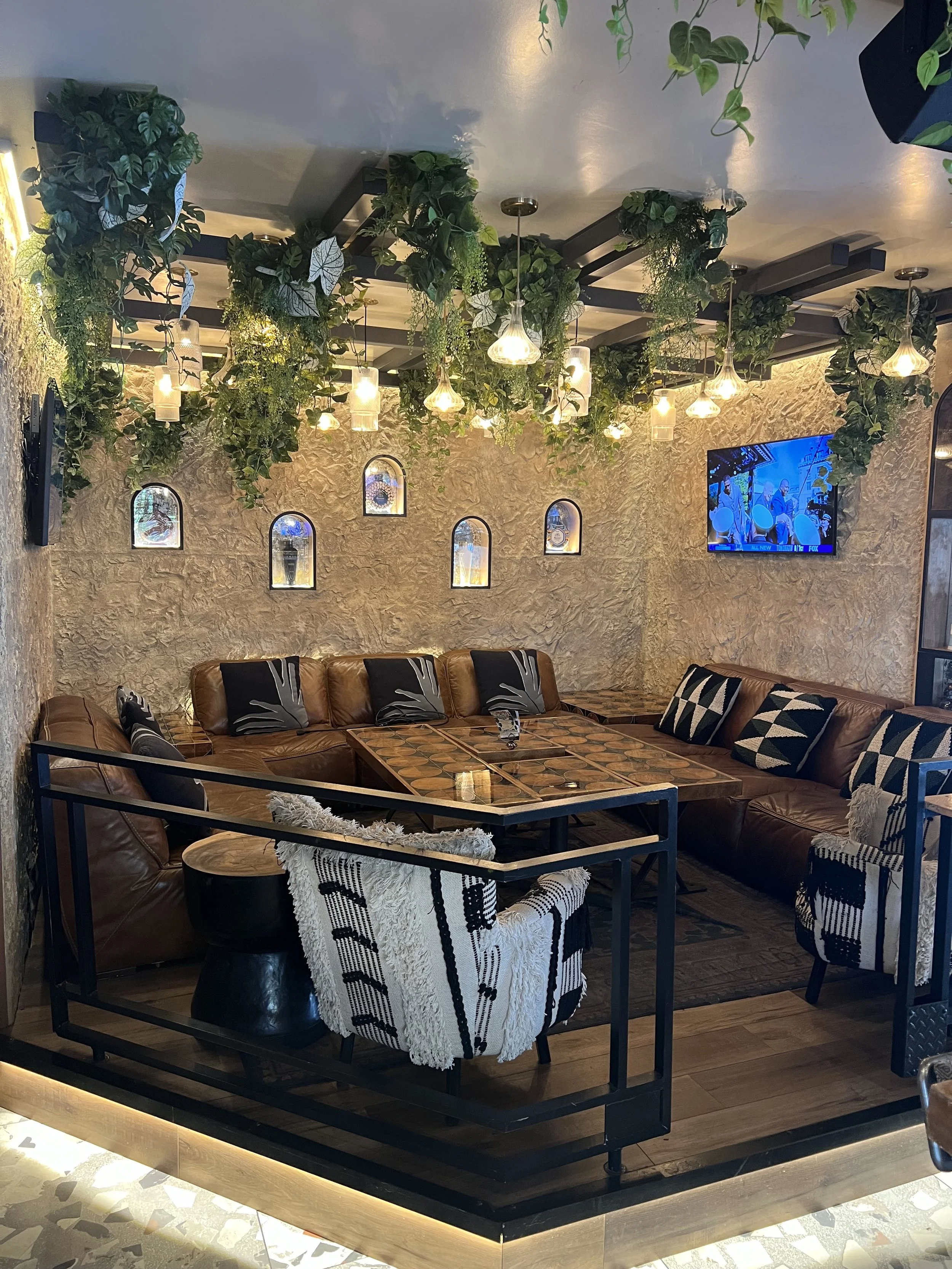 Interior of a cozy lounge area with a leather sectional sofa, patterned pillows, a wooden coffee table, decorative wall niches with art, greenery on the ceiling, and a wall-mounted TV.