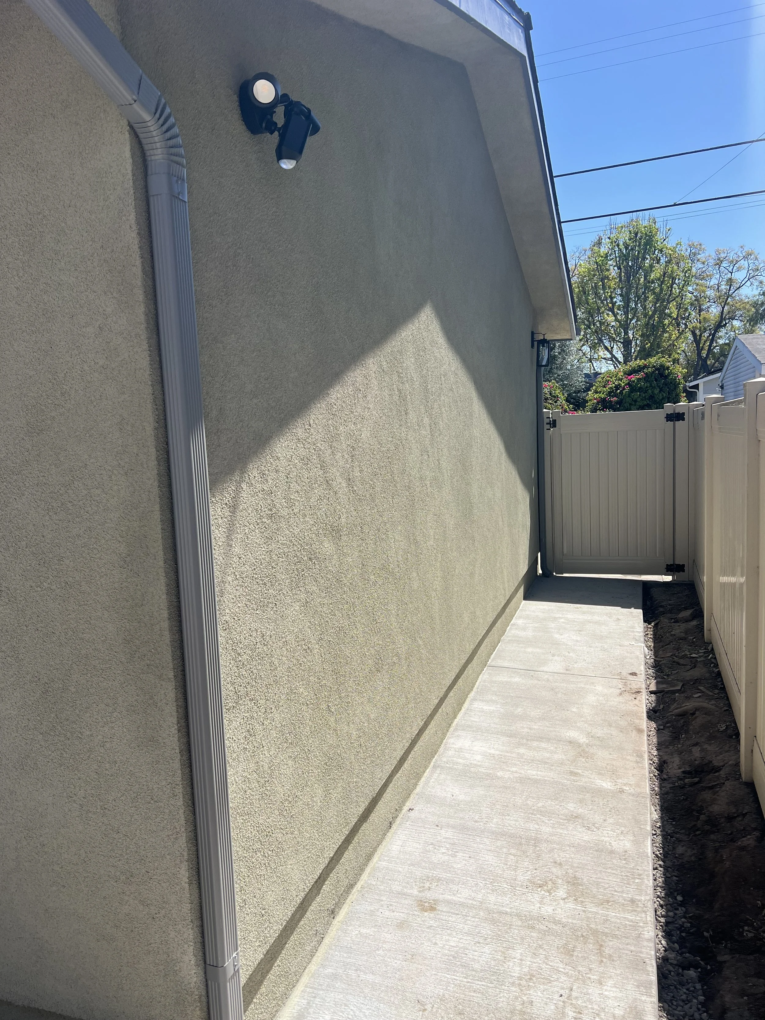 The image shows a beige exterior wall of a house with a mounted outdoor light fixture. A section of a white fence and gate is visible in the background, with some trees and bushes behind the fence. There is a concrete walkway along the base of the wa