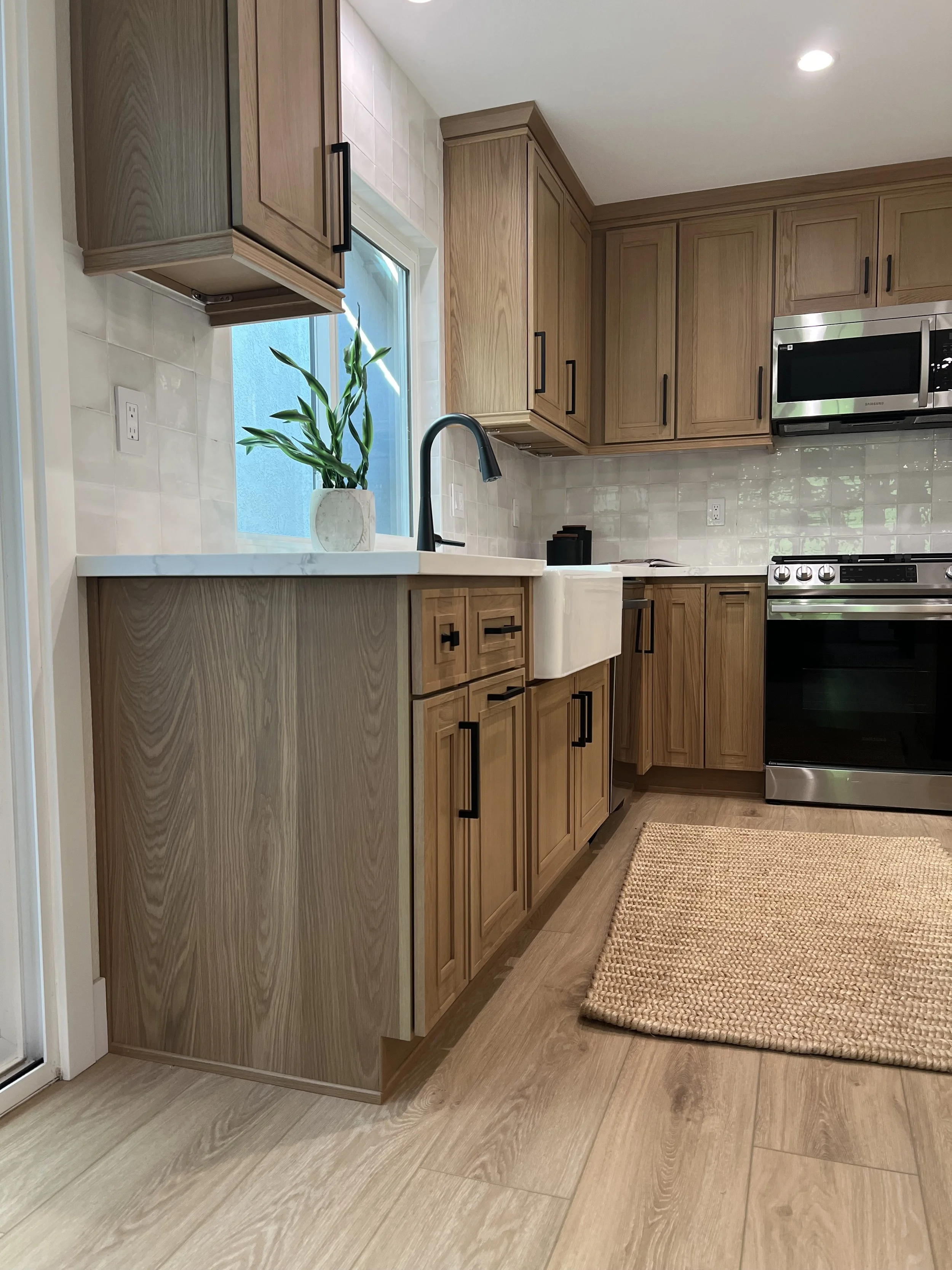 A modern kitchen with wooden cabinets, a black sink faucet, a white farmhouse sink, a potted plant on the counter, a stainless steel microwave, a stove, a beige rug, and a window with a blue exterior wall visible outside.