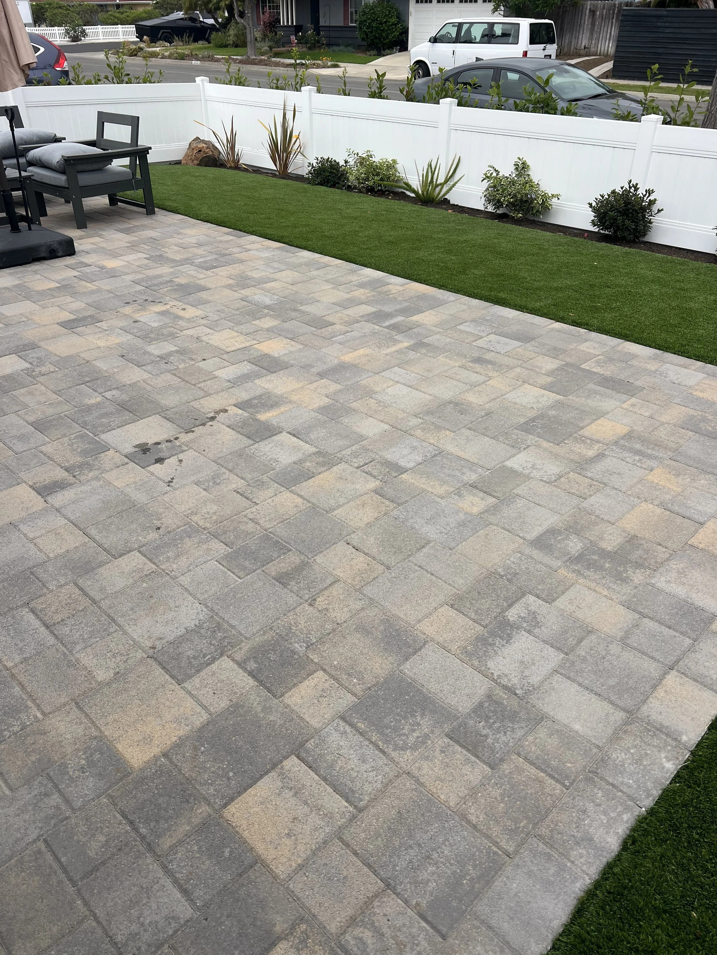 A backyard patio area with a brick paver surface, a white fence, and features including outdoor seating, plants, and a manicured lawn.