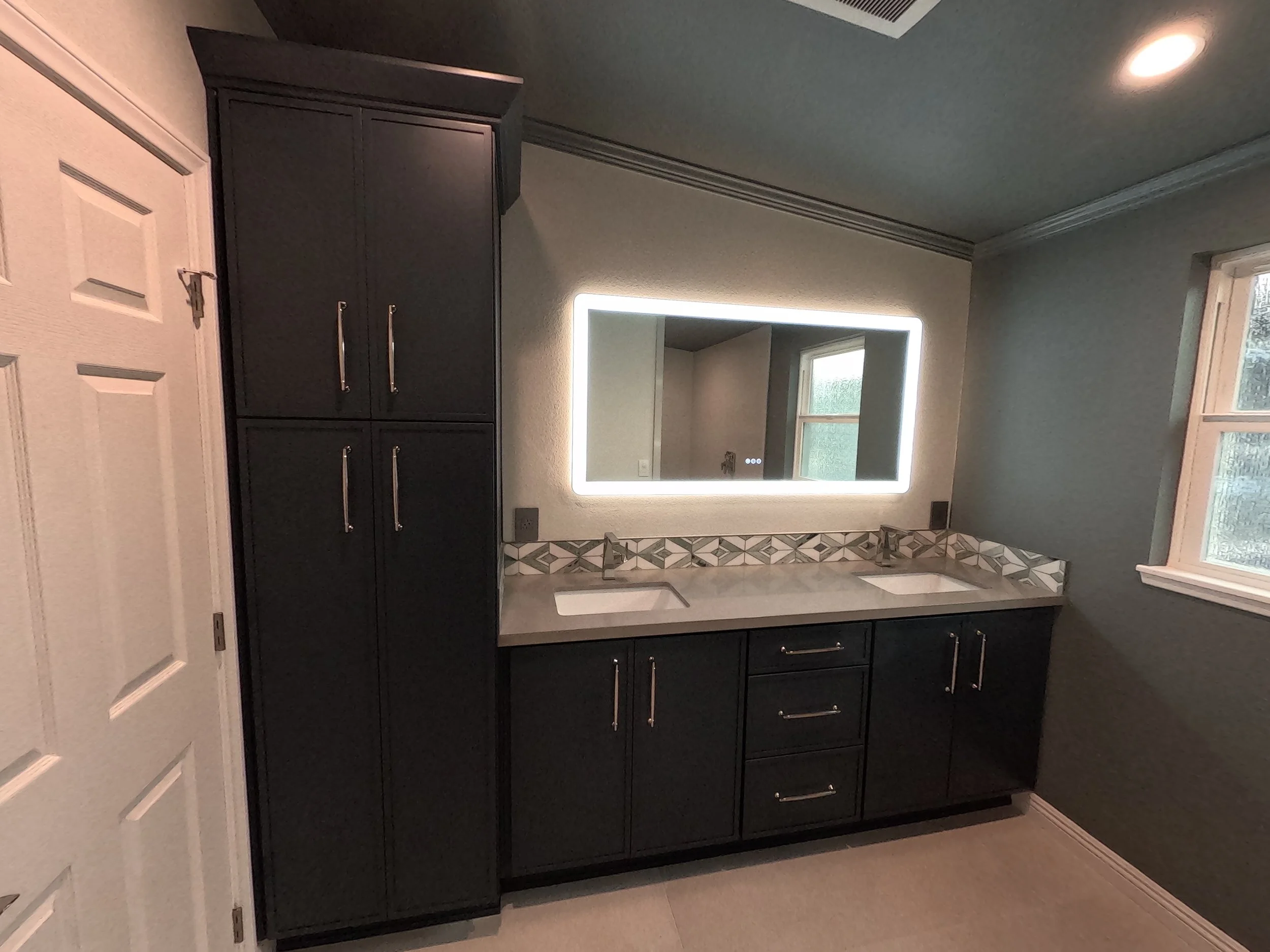 Bathroom vanity with black cabinets, white countertop, large illuminated mirror, and two small windows.