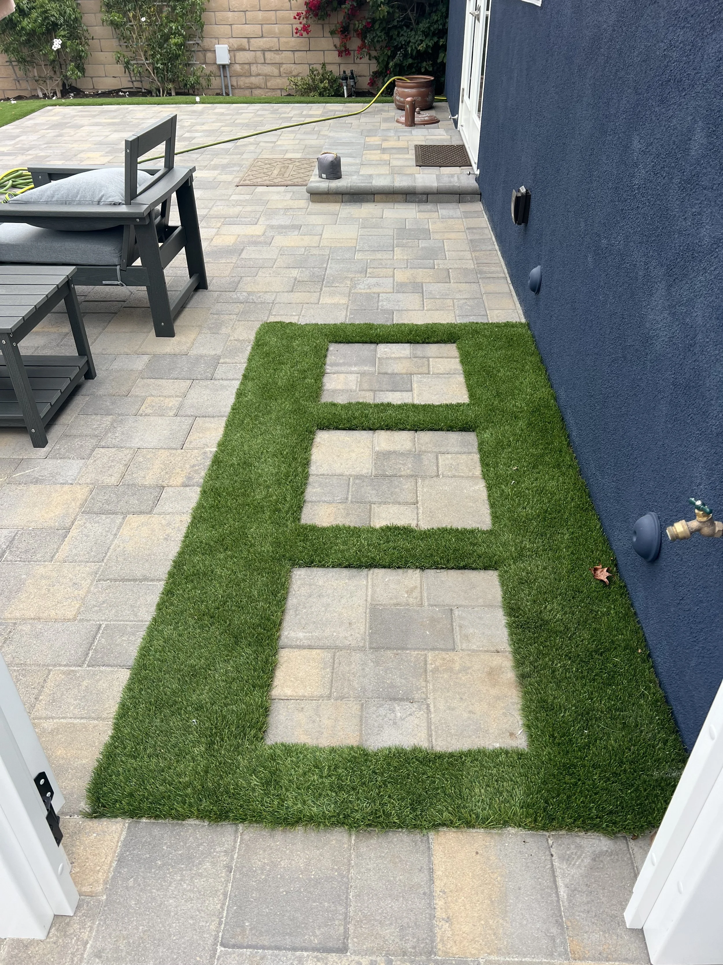 A patio with paving stones, a blue house wall, a small patch of artificial grass with square cutouts, and a pathway leading to an entrance with stairs. There is a bench with a pillow, a garden hose, a mat, and some plants and flowers in the backgroun