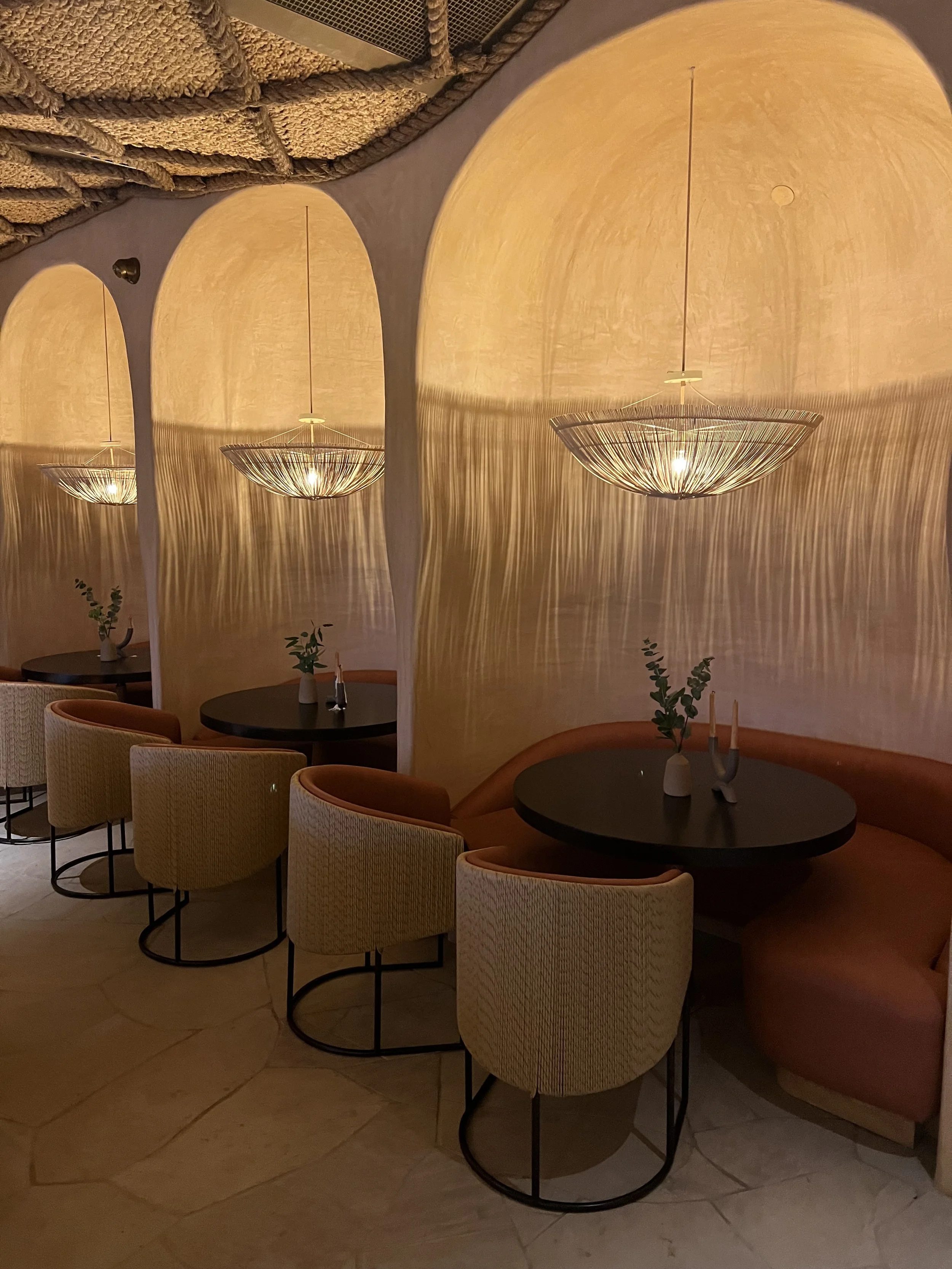A dimly lit dining area with several beige upholstered chairs around small round black tables. Vases with green leafy plants and candles are on the tables. The wall has a unique textured appearance with large semi-circular light fixtures creating pat