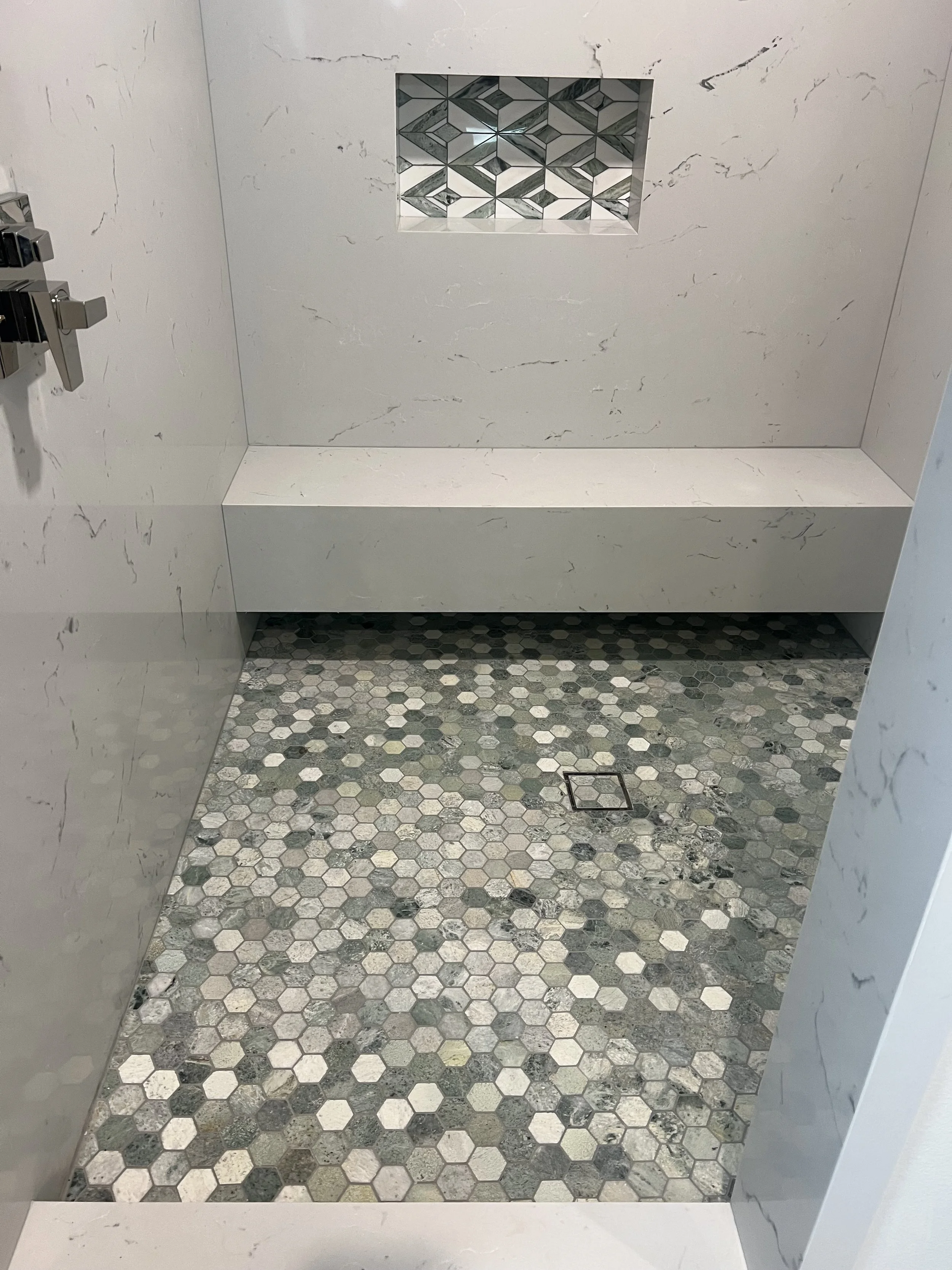 A shower area with white marble walls and a hexagon tile floor in shades of gray, white, and black. There is a square drain on the floor and a decorative niche with geometric patterned tiles in the wall.