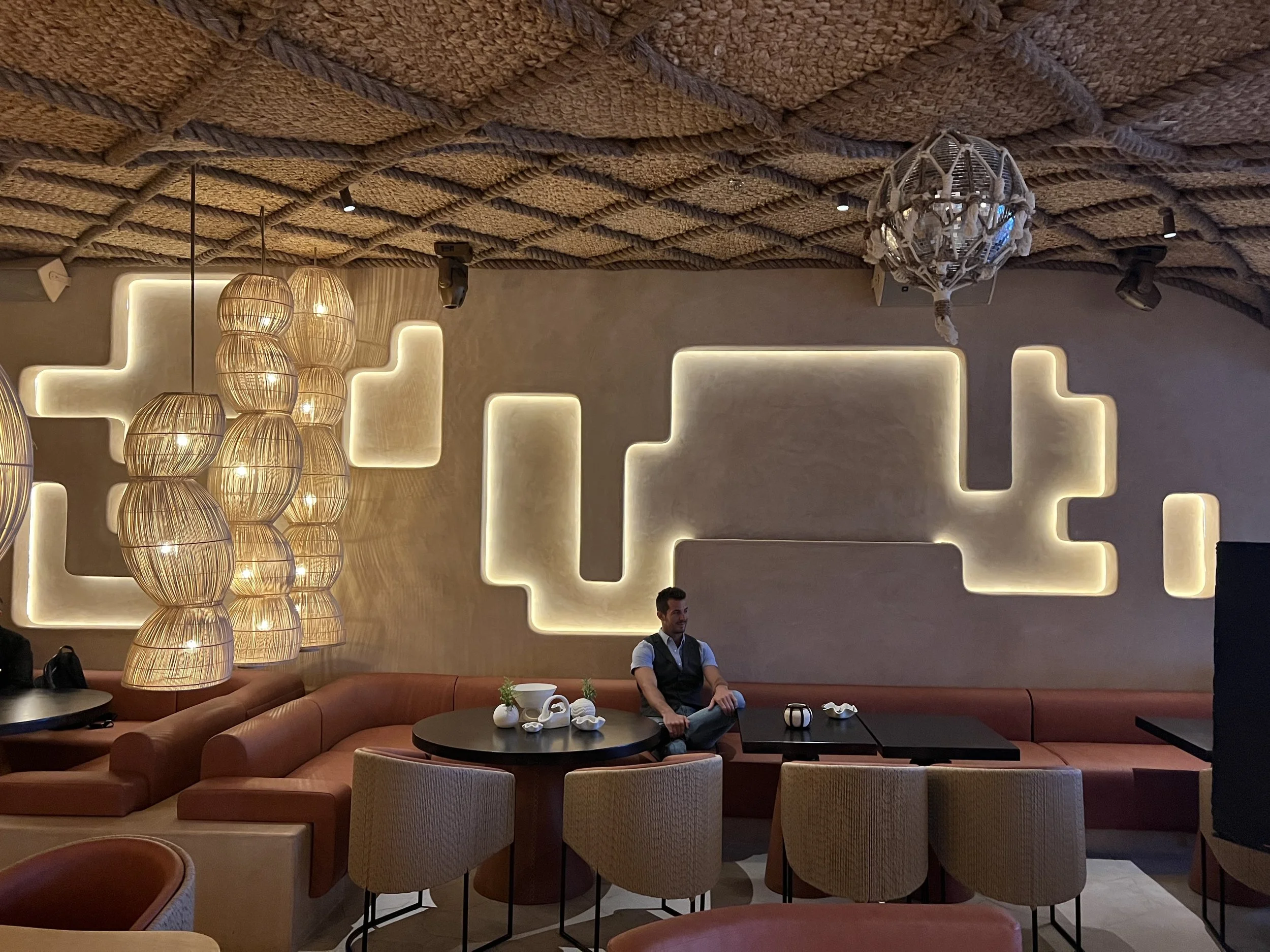 A modern hotel lobby with geometric wall design, decorative lighting fixtures, a curved sectional sofa, a table with small decorative items, and a man sitting on the sofa.