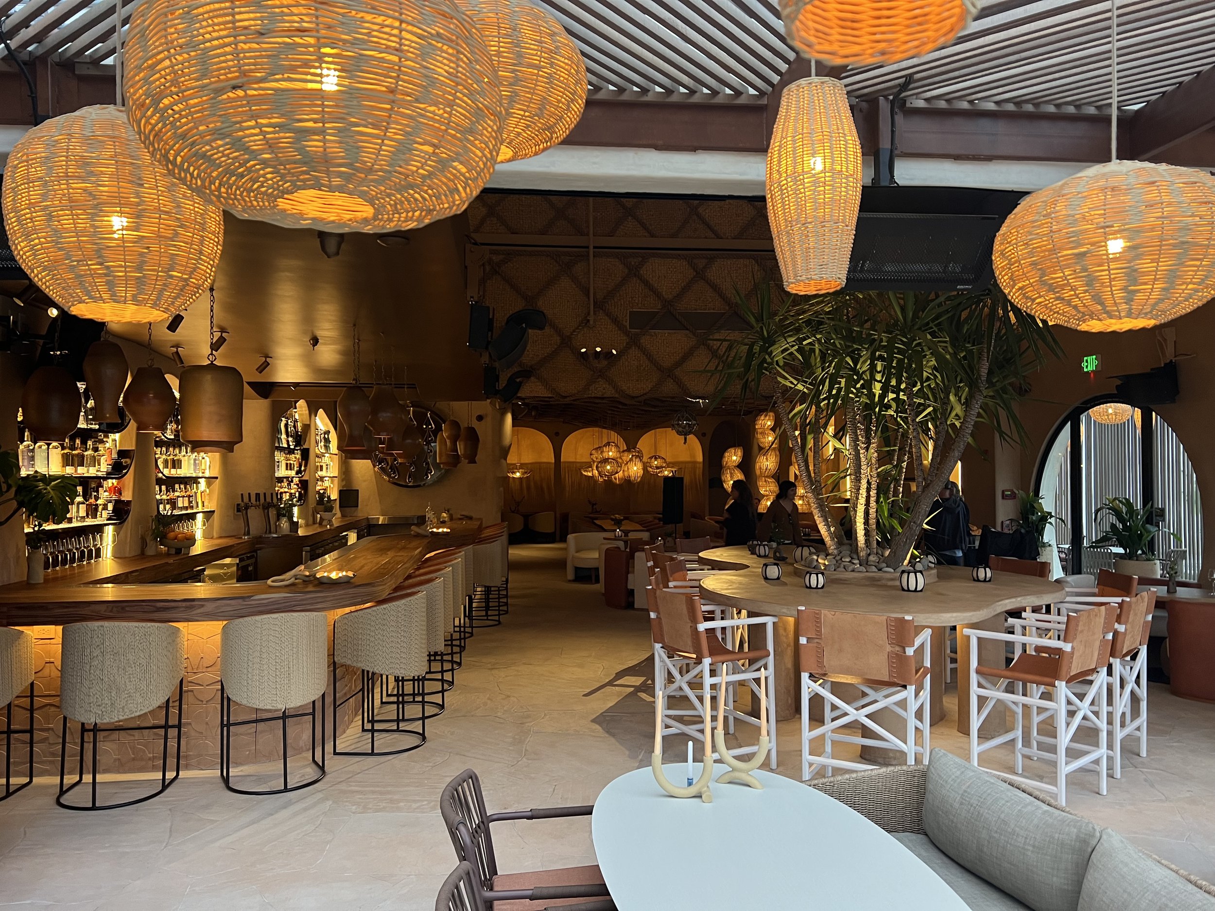 Interior of a restaurant or bar with hanging woven lamps, a curved bar counter with barstools, a tree in the center, and several tables and chairs for seating.