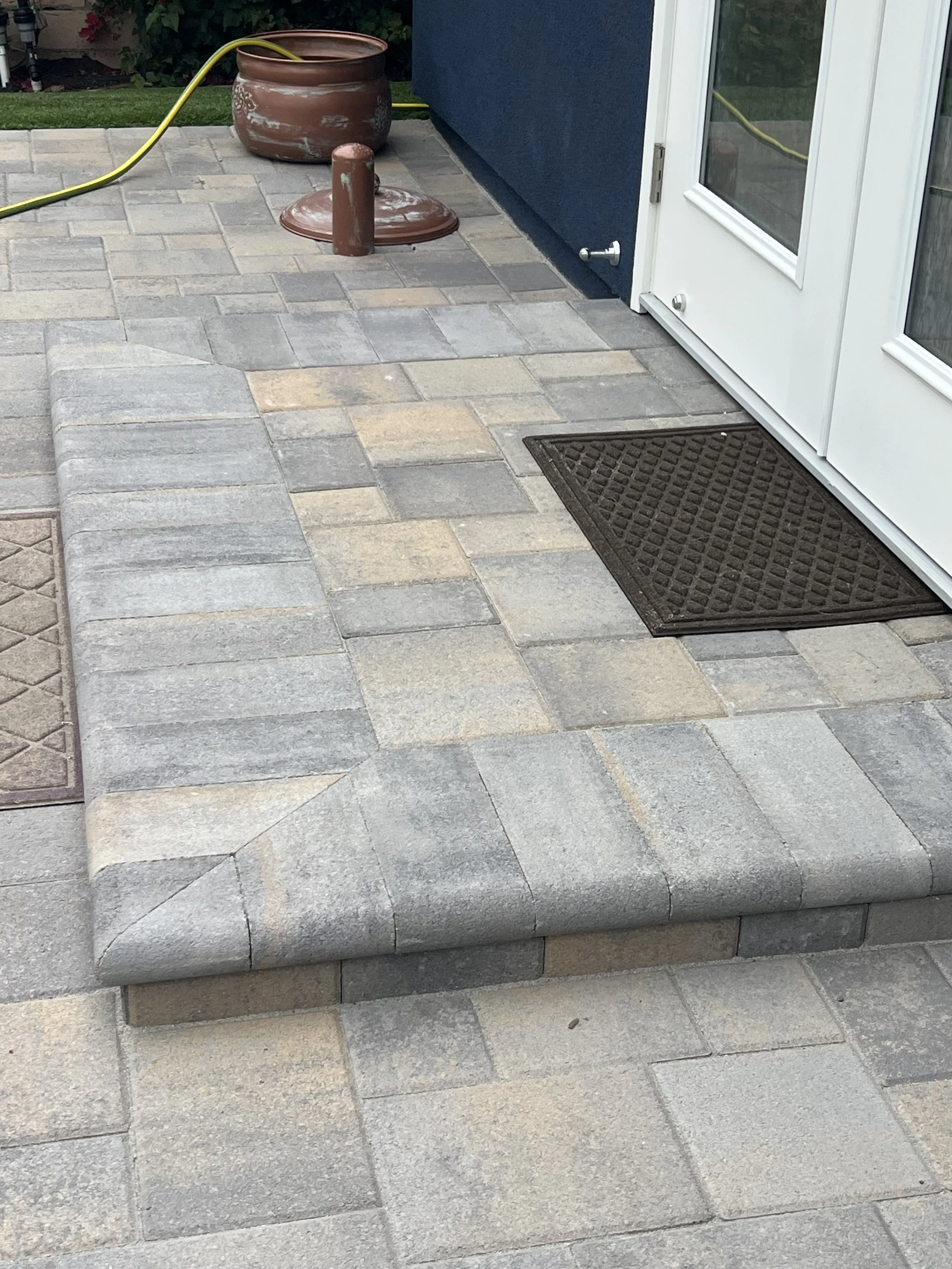 Paved patio with stacked stone steps and a dark brown doormat near a white door with glass panes. A large plant pot with a yellow garden hose, a brown cover, and a post are in the background.