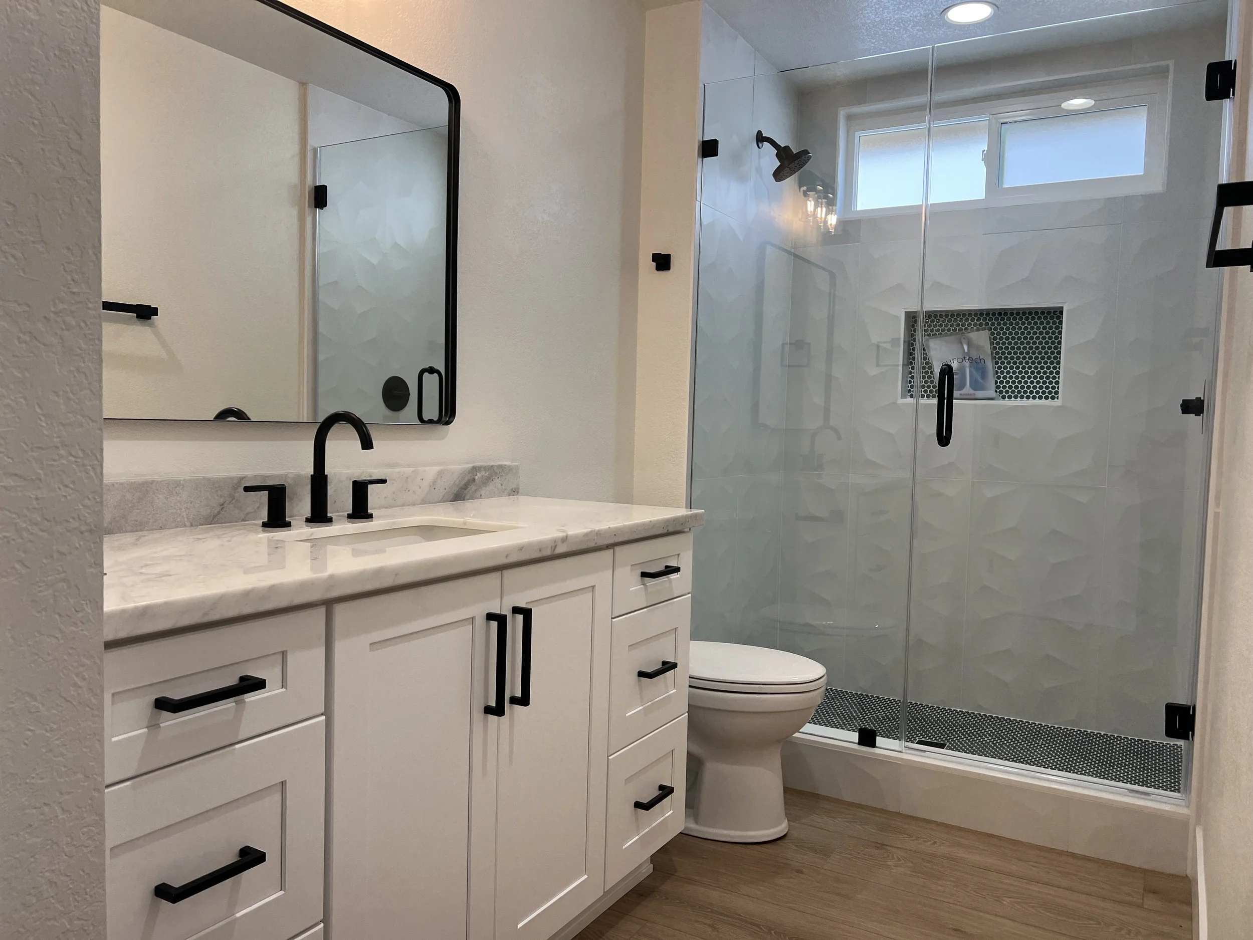 Modern bathroom with white vanity, black hardware, and marble countertop. Includes a toilet and a glass shower with black fixtures and a window.