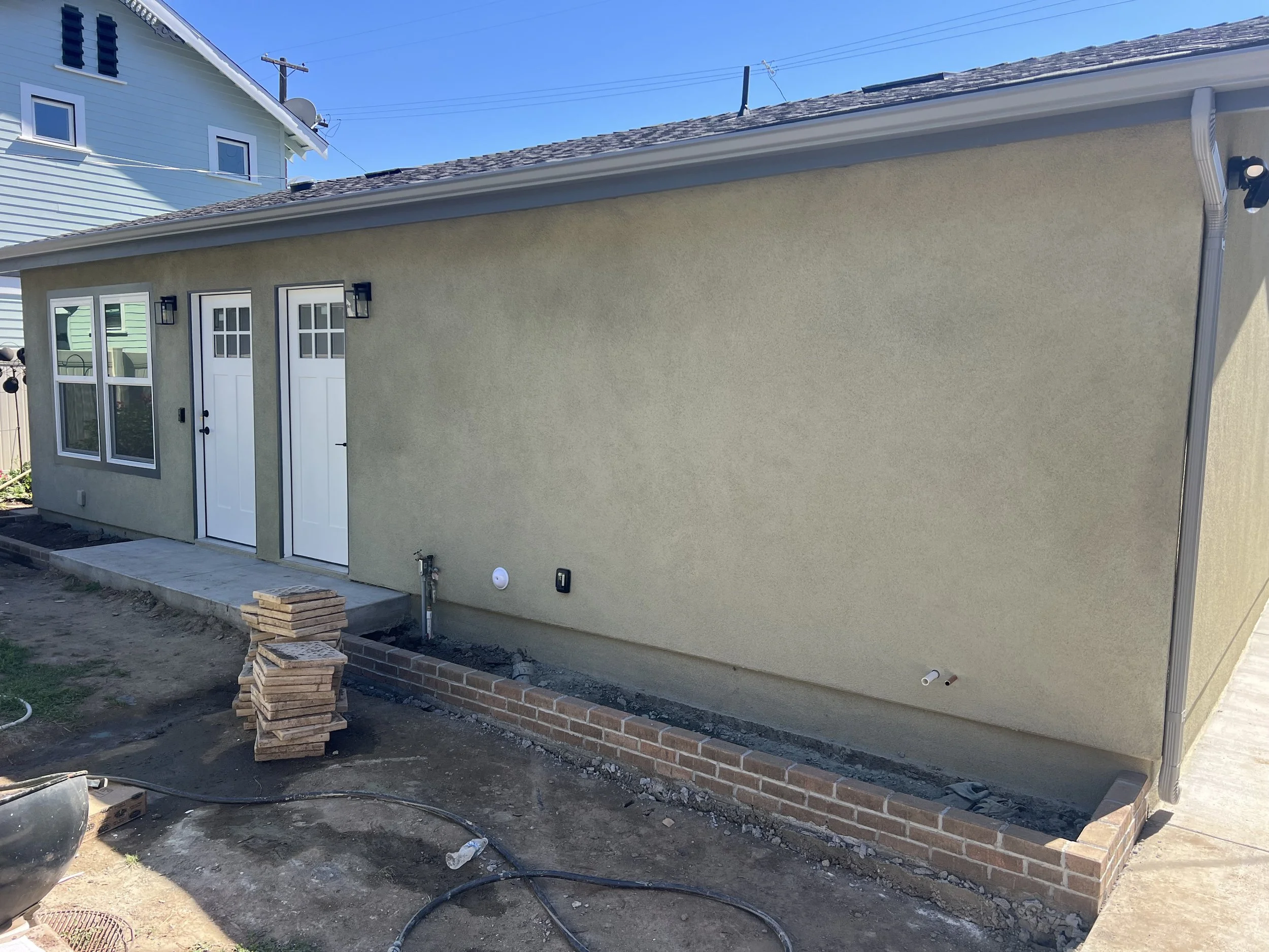 Backyard with construction work in progress. There are two white doors with glass panels, a window with multiple panes, and outdoor lights attached to the house's wall. A small brick wall is being built along the base of the house, and construction m