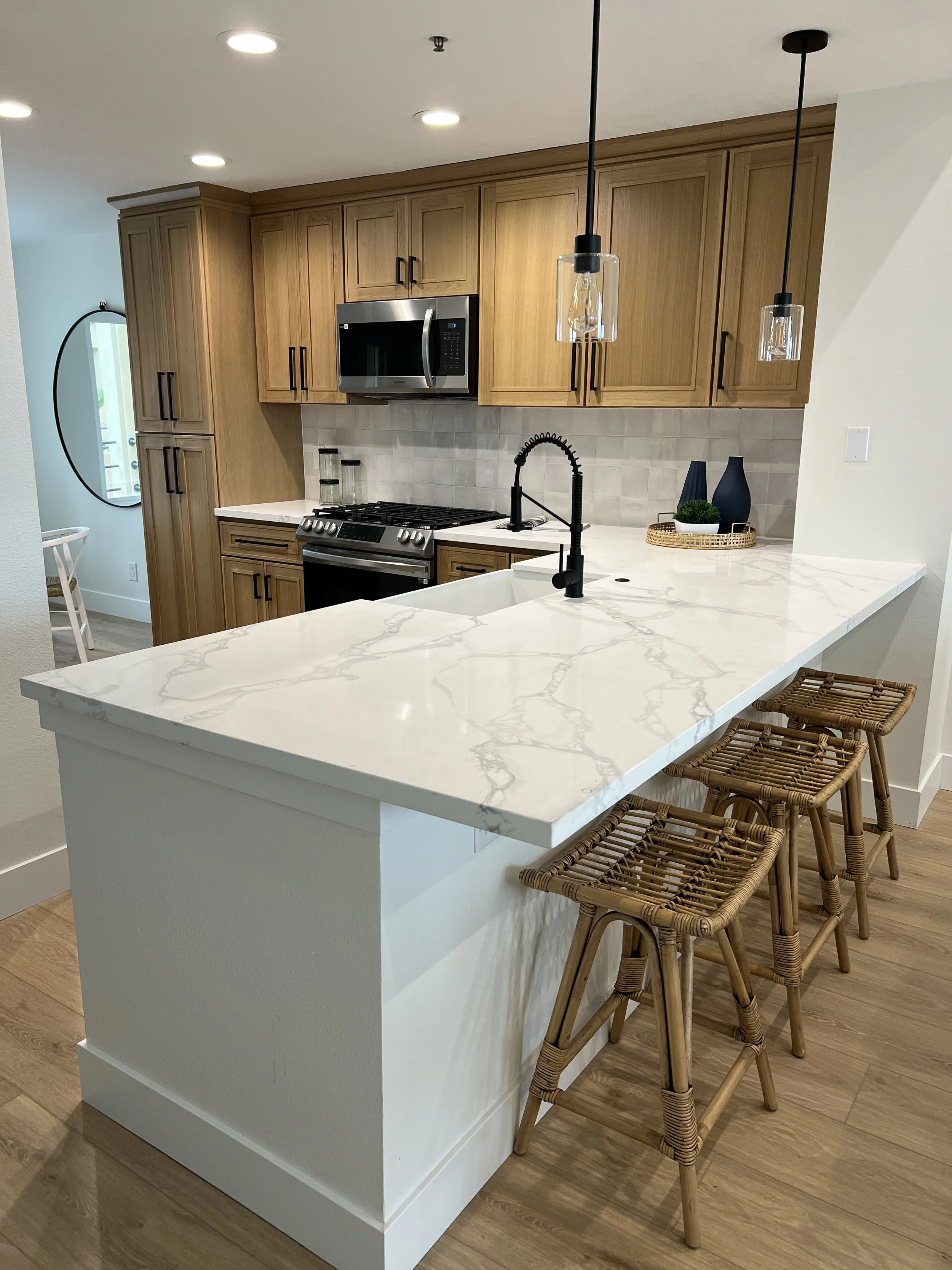 Modern kitchen with wooden cabinets, white marble countertop, black sink faucet, stainless steel microwave and stove, decorative vases, and a small tray of plants. Pendant lights hang above the counter, and wooden bar stools are lined up in front.