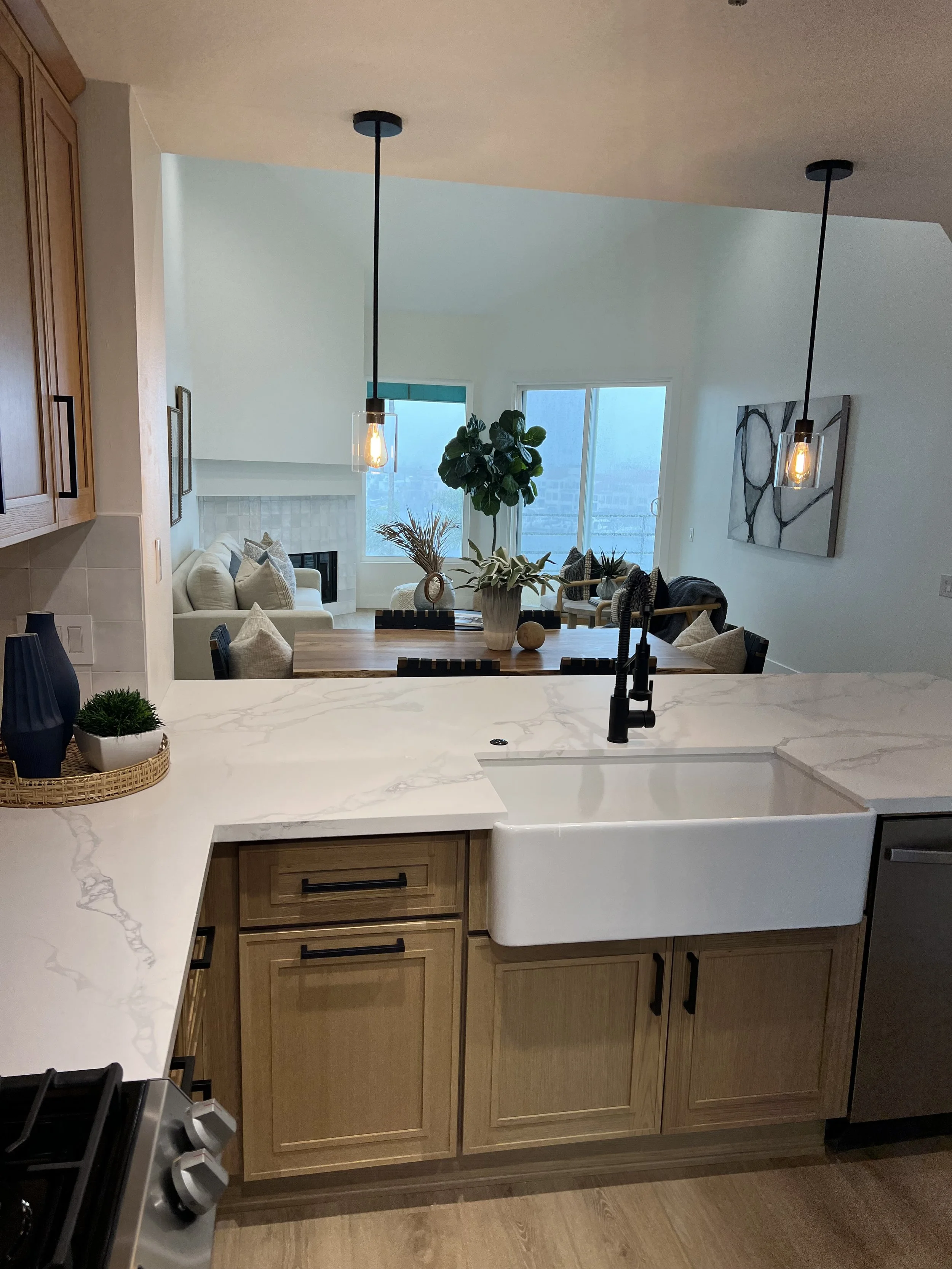 A modern kitchen with a white marble countertop, wooden cabinetry, black hardware, and a black faucet. In the background, there is a dining table with potted plants and decorative items, and a living room with a sofa and large windows.