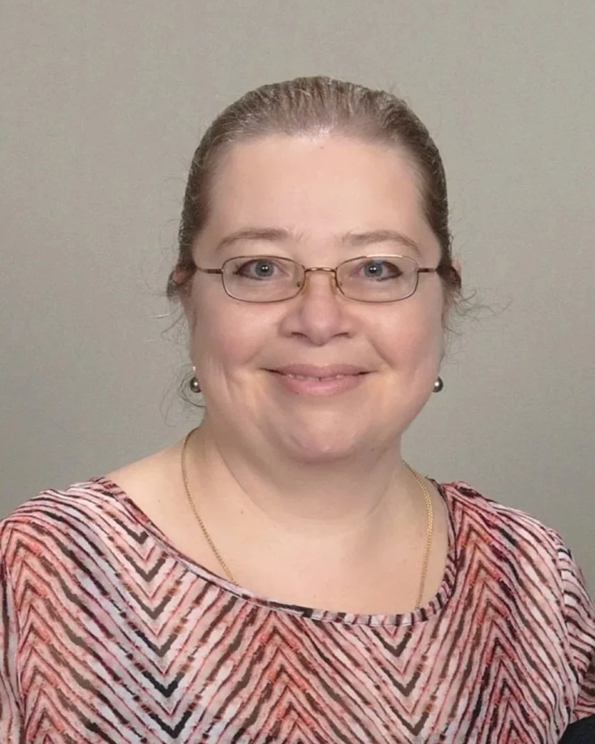 A woman with glasses smiling at the camera, wearing a patterned top and jewelry, against a plain background.