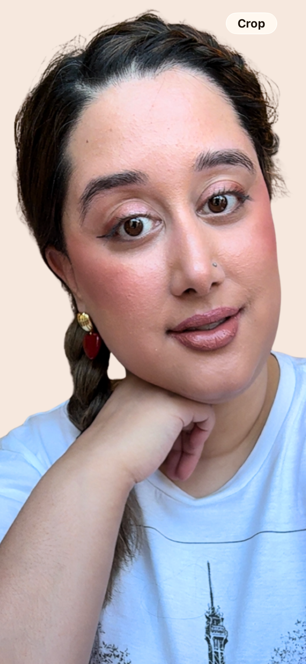 Close-up of a woman with dark hair, wearing makeup, earrings, and a light blue T-shirt with a graphic design, resting her chin on her hand against a plain background.