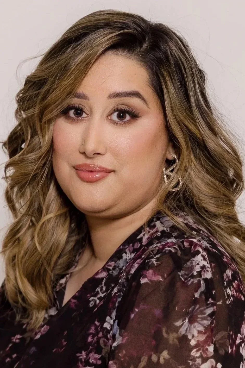 A woman with long, wavy hair, wearing makeup, earrings, and a floral patterned top, smiling softly against a plain background.