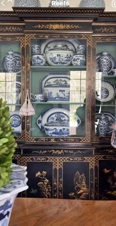 Wooden display cabinet with blue and white Chinese porcelain dishes and vases inside, decorative gold accents, and a green background.