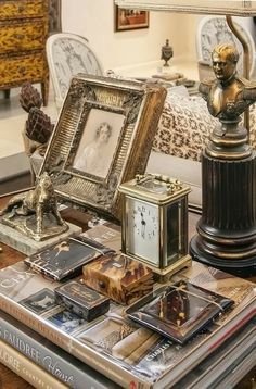 A collection of vintage and antique items on a glass table, including picture frames, a clock, a bust sculpture, and several decorative books, in a decorated room.