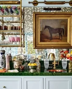 A kitchen with a green countertop, gold fixtures, and a framed painting of a horse on the wall. Shelves with pink and clear cups and drinking glasses are also visible.
