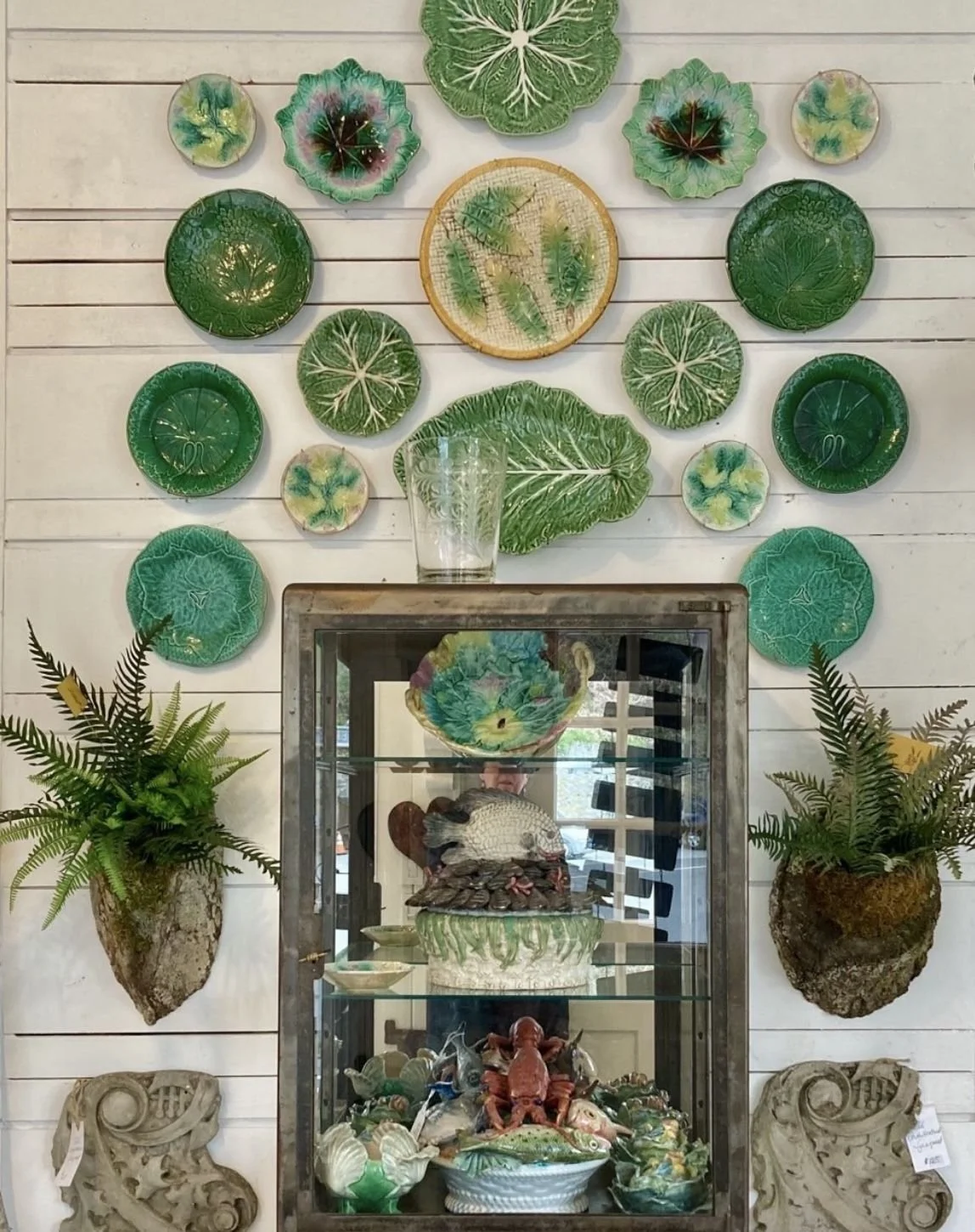 Display of decorative green ceramic plates and bowls with leaf patterns mounted on a white wooden wall, with potted ferns on either side and a glass display case in the center filled with various ceramic sea life and shell sculptures.