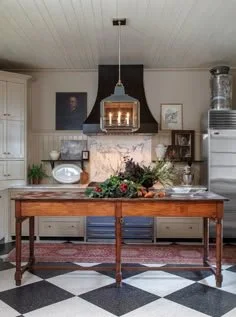 A kitchen with a wooden table, white cabinets, a black range hood, and various plants and decorations. The floor has a black and white checkered pattern.